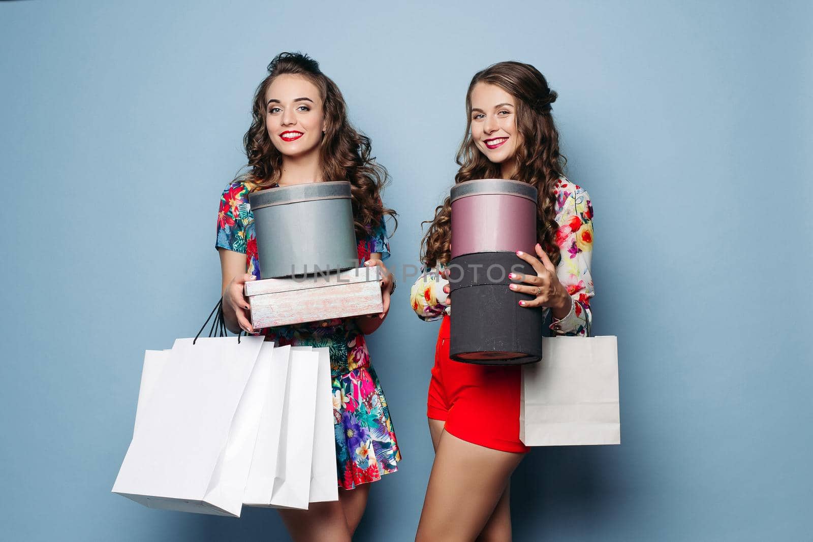 Portrait of beautiful brunette ladies with hairstyles in trendy clothes holding hat bags and shopping bags over blue backgroud. Smiling at camera.