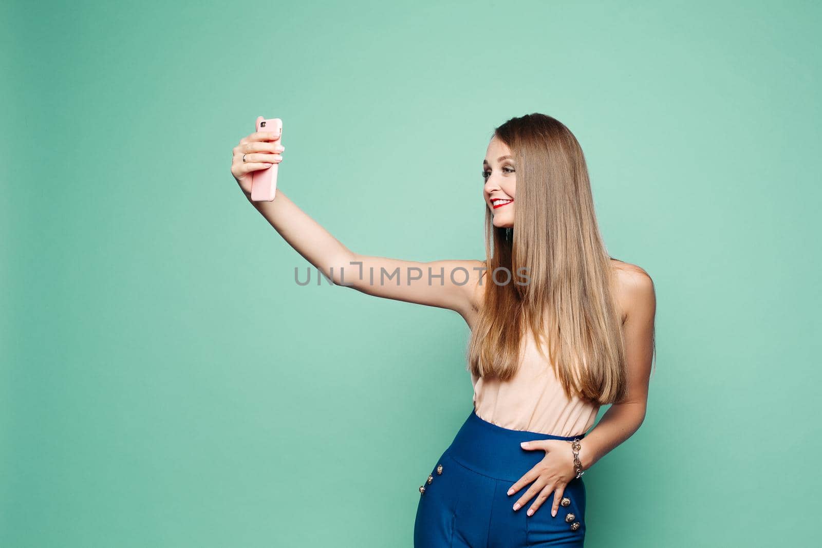 Studio portrait of beautiful elegant blonde woman with straight hair in blue trousers and top taking selfie with mobile phone over green background. Isolate.