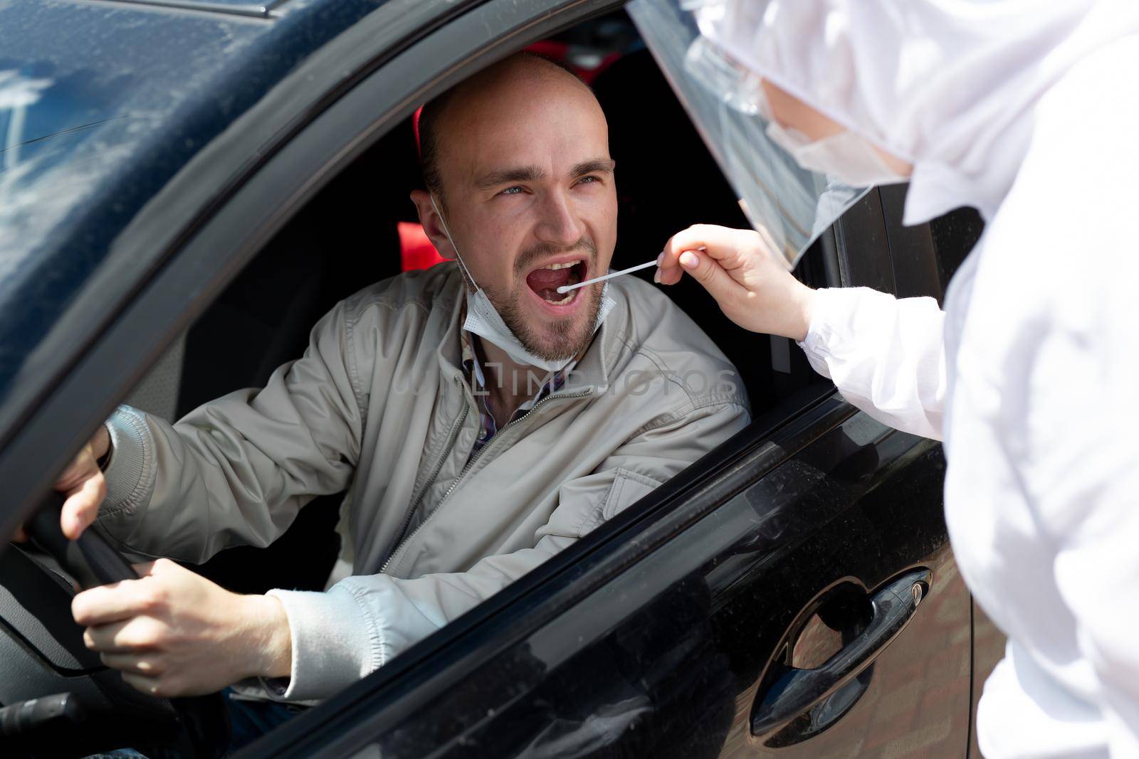 A man wearing a protective suit, gloves, surgical mask, and face mask is testing the coronavirus co id-19 on another man sitting in a car.