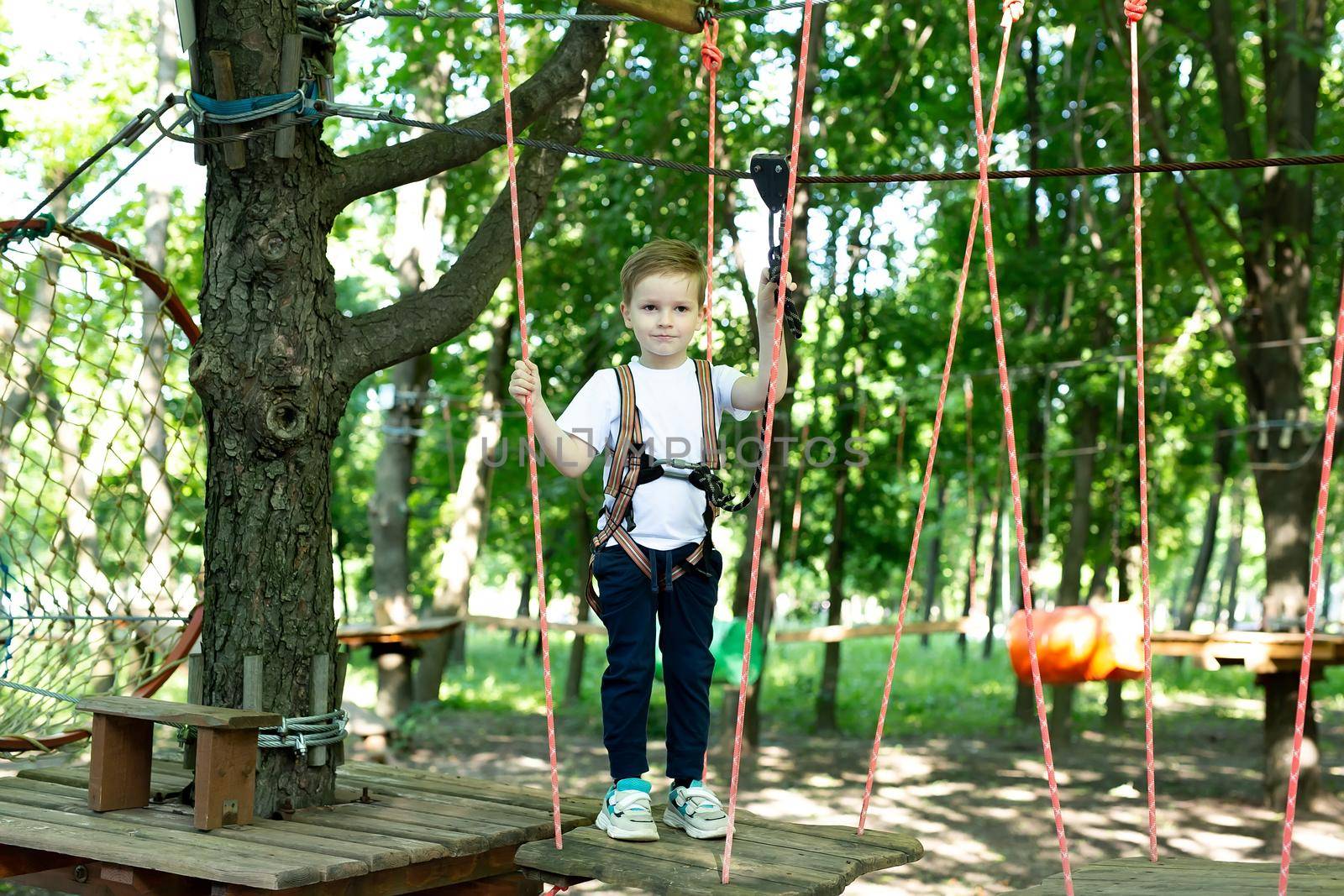 Small boy in climbing gear is walking along a rope road in an adventure Park, holding on to a rope and a carabiner by StudioPeace