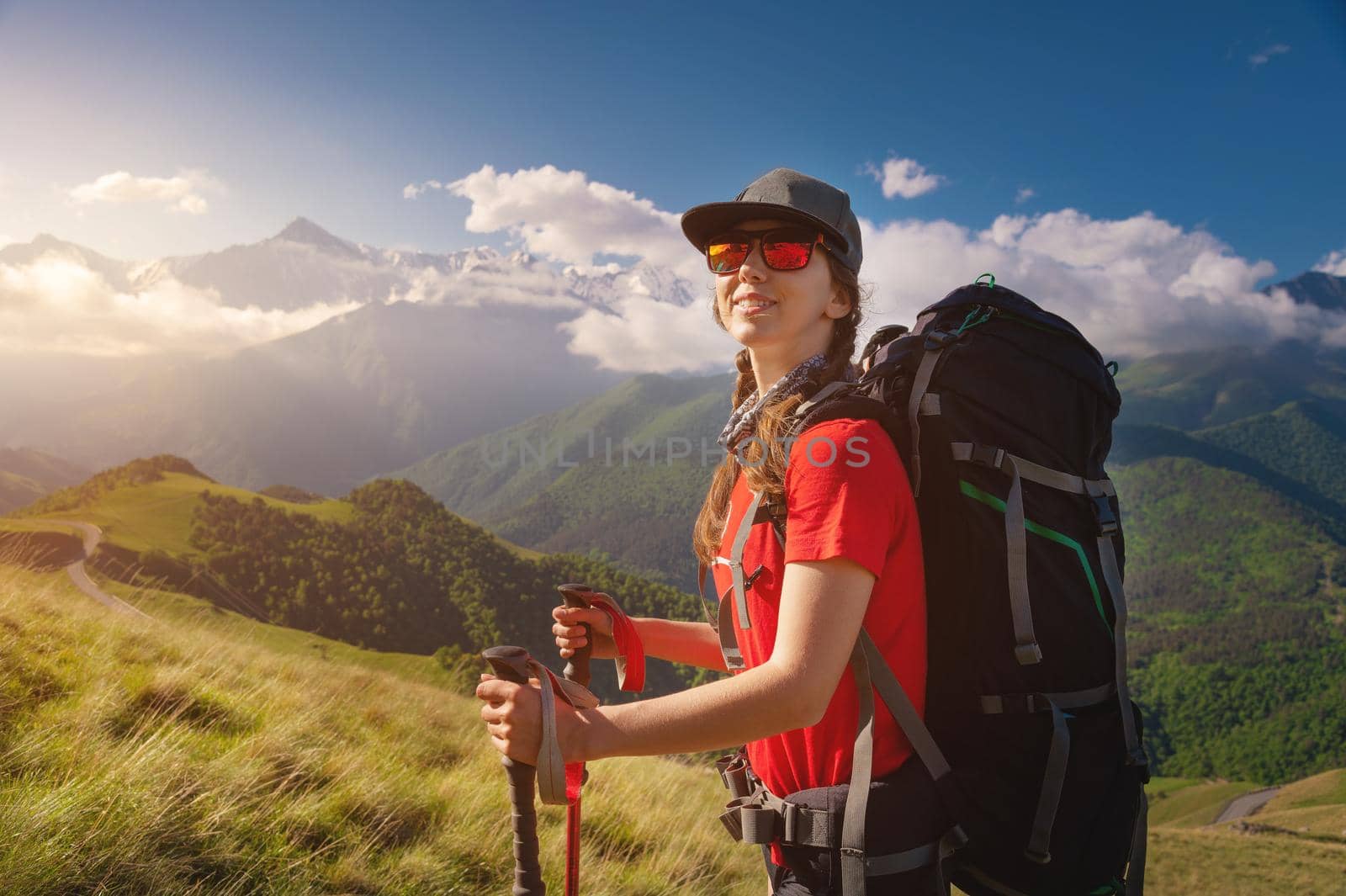 Caucasian traveler in a cap and sunglasses with a backpack on her back and trekking poles against the backdrop of high mountains and low clouds. Smiling and looking at the camera.