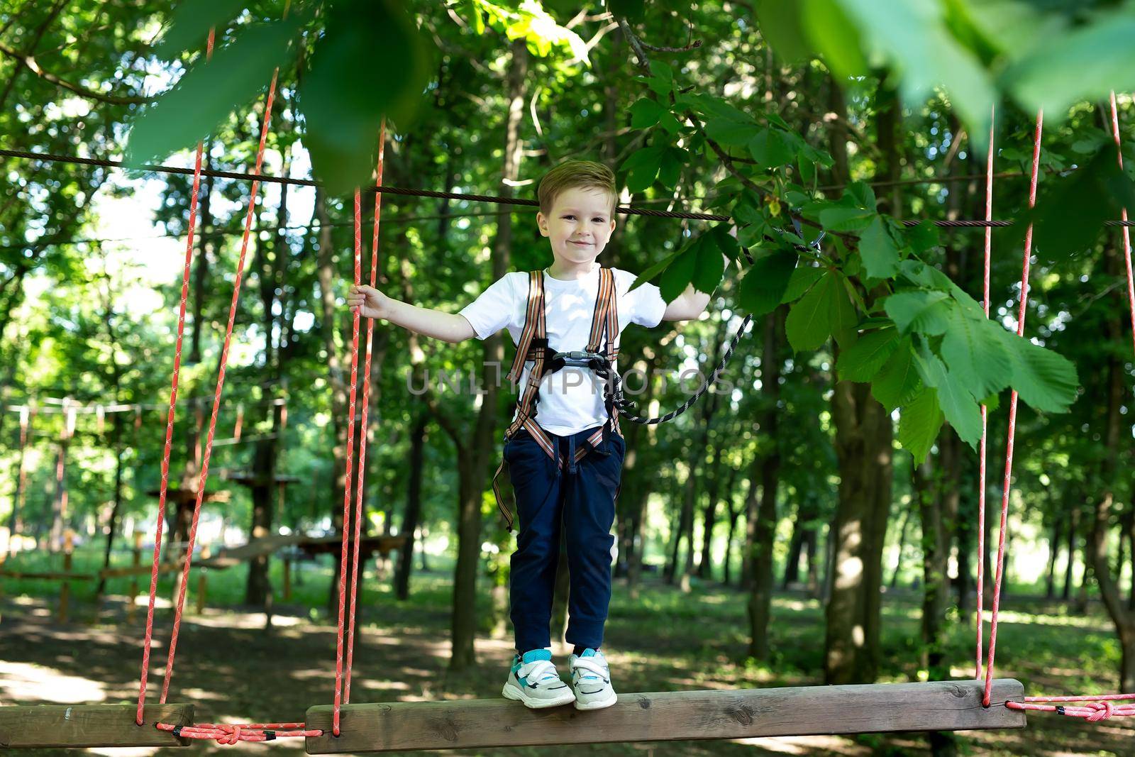 Small boy in climbing gear is walking along a rope road in an adventure Park, holding on to a rope and a carabiner by StudioPeace