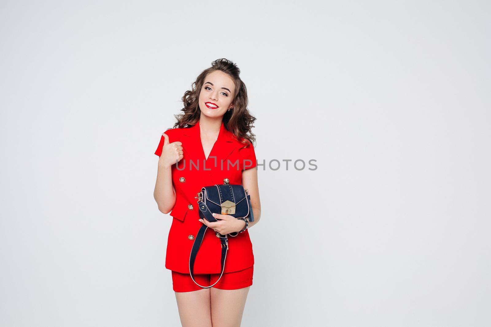 Studio portrait of attractive smiling woman with hairstyle and make up wearing bright red suit and holding trendy blue leather crossbag, showing thumb up with toothy smile. Isolated on white.
