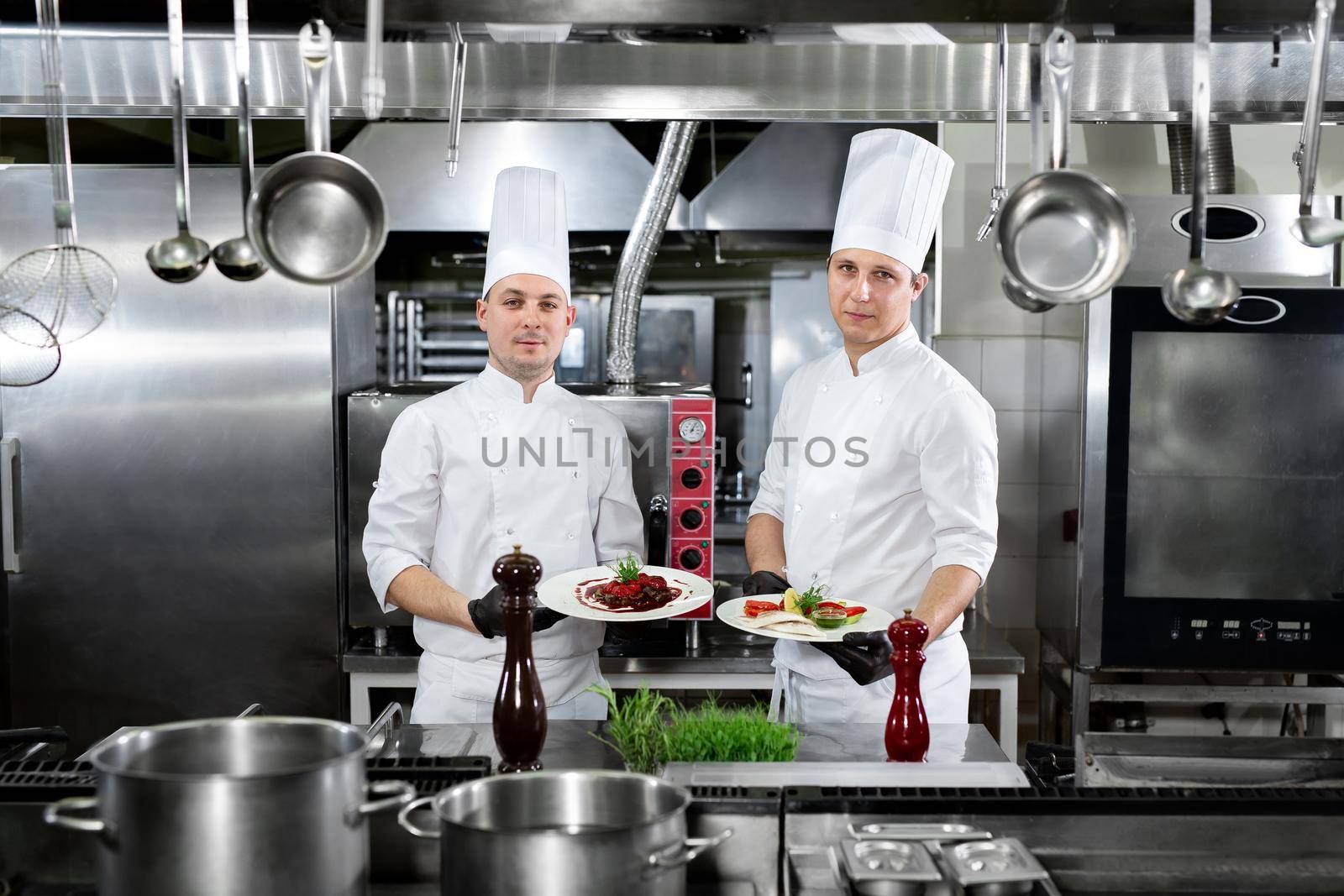 Two chefs in a professional kitchen hold ready-made dishes in their hands