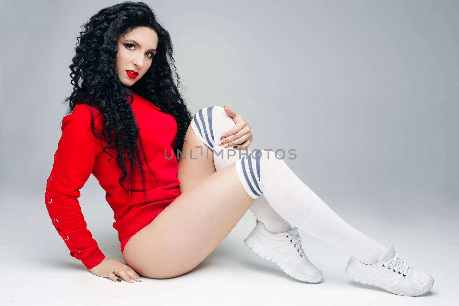 Portrat of sexy and attractive fit young brunette in red, sitting and looking at camera. Swag stylish model with curls long hair and red lips posing and touching leg. Concept of lifestyle sport.