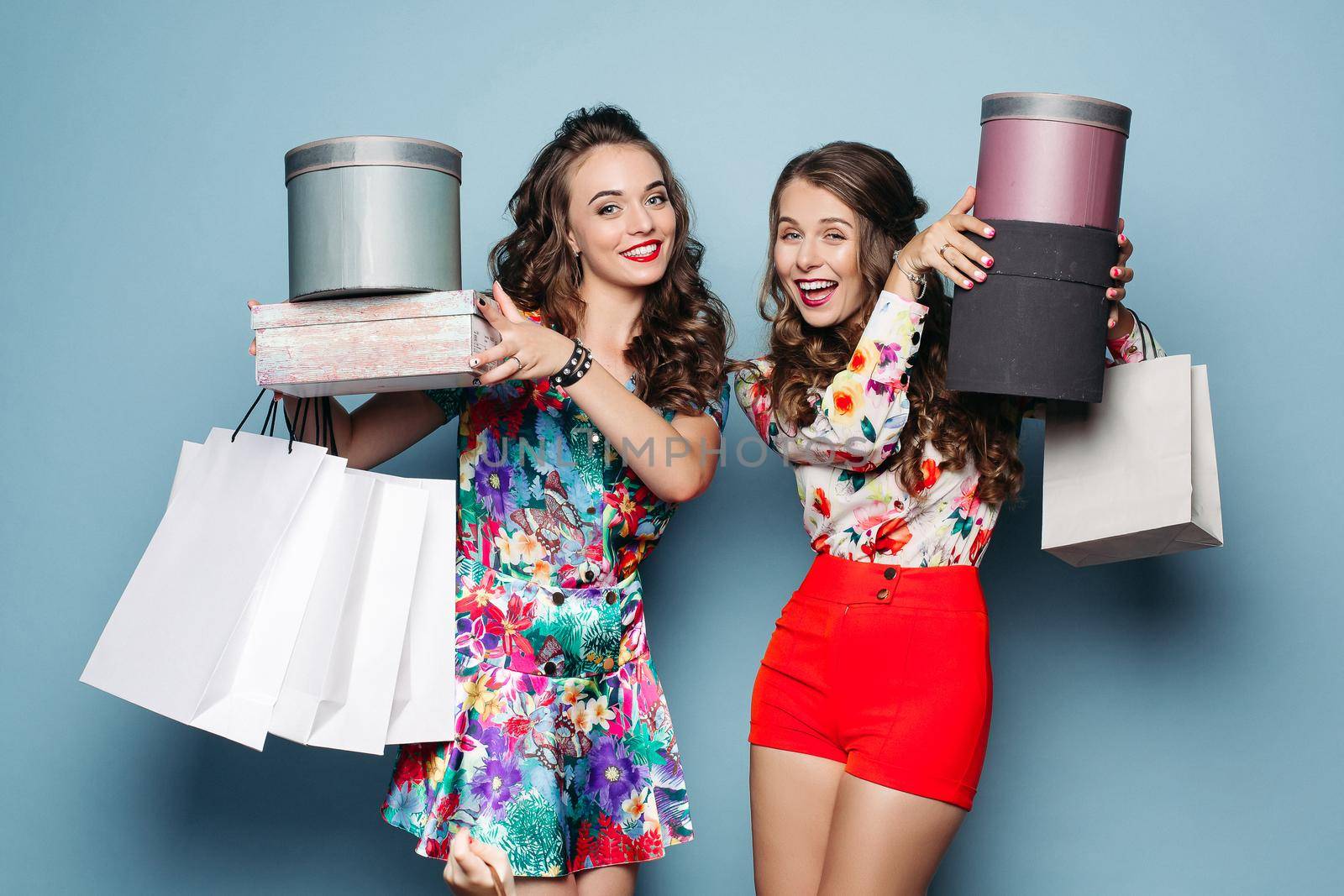 Studio portrait of happy smiling two best girlfriends in colorful fashionable clothes after shopping. Women holding hands up with many paper bags, boxes, presents. Concept of deason sale and shopping.