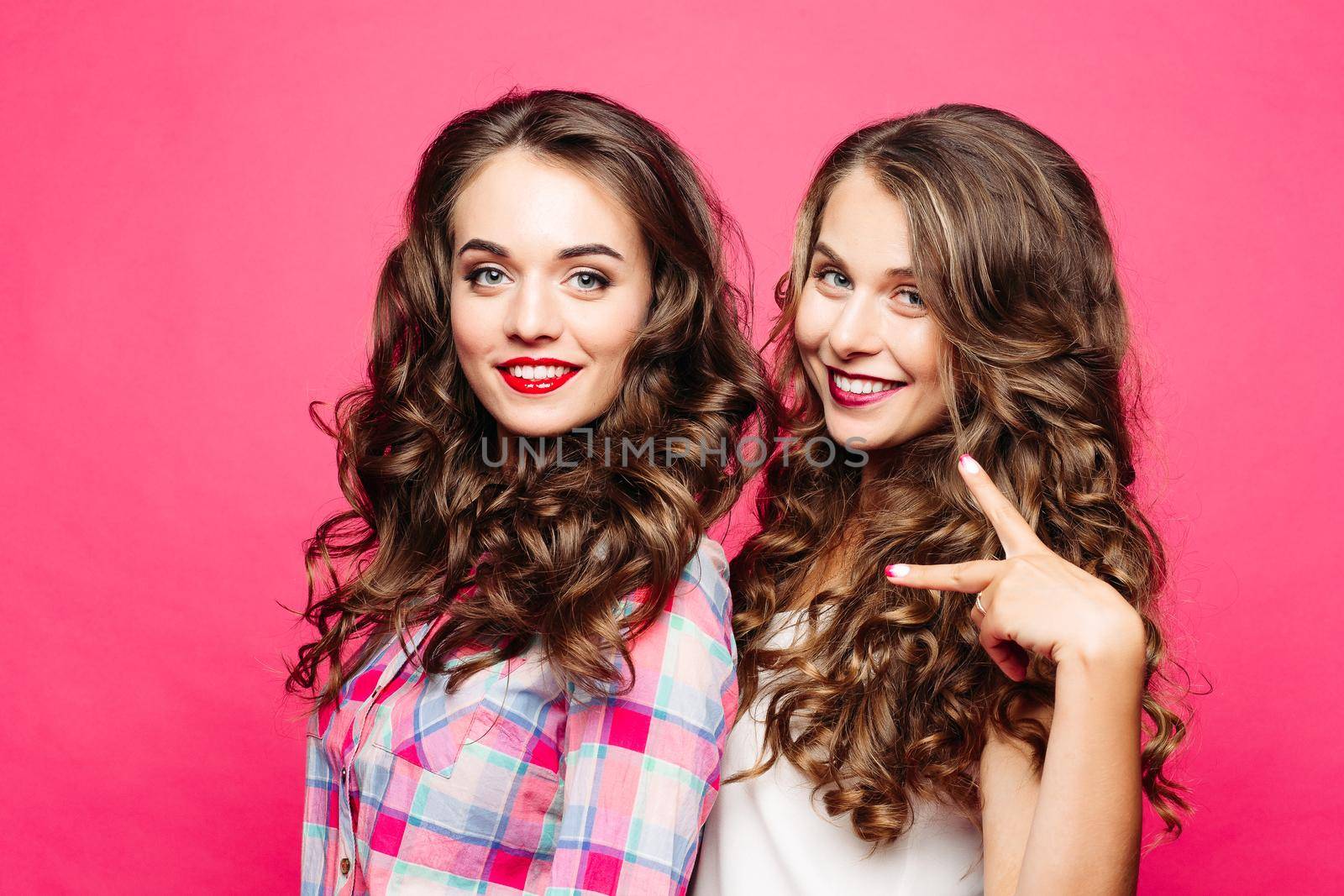 Portrait of stunning beauties with gorgeous wavy hair and red lips smiling at camera and showing peace sign. Isolate on pink background.