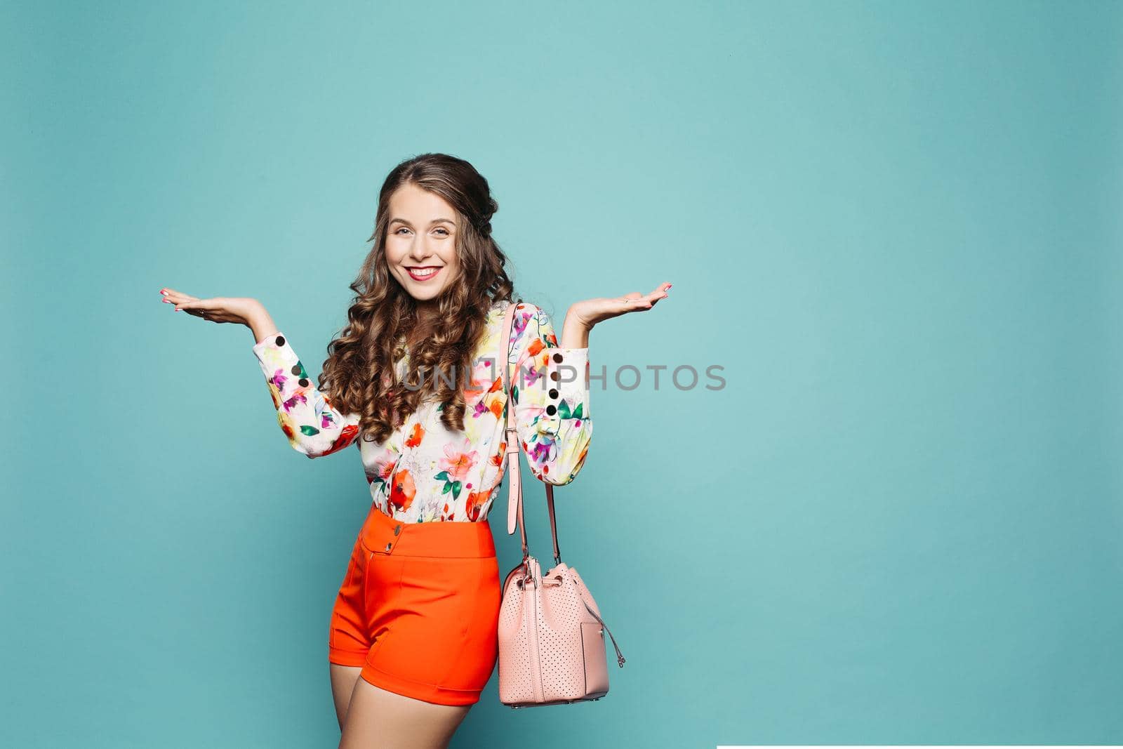 Studio portrait of attractive woman with curly hair wearing floral shirt with red shorts and carrying bag demonstrating two objects over green background. Isolate.