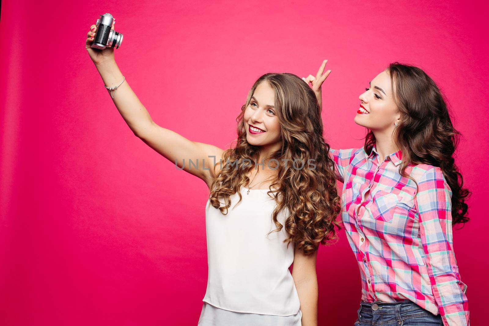 Portrait of happy attractive girlfriends with long wavy hairstyles taking self-portrait via old film camera against red background. Brunette in plaid shirt making bunny ears to her friend.