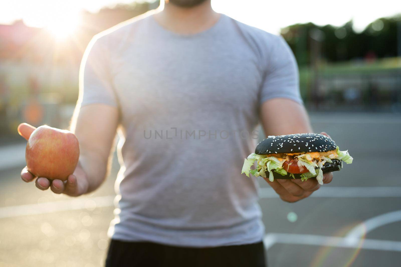 Young athletic man in a t-shirt holds a Burger and an Apple in the background of the stadium, a choice of healthy and unhealthy food. by StudioPeace