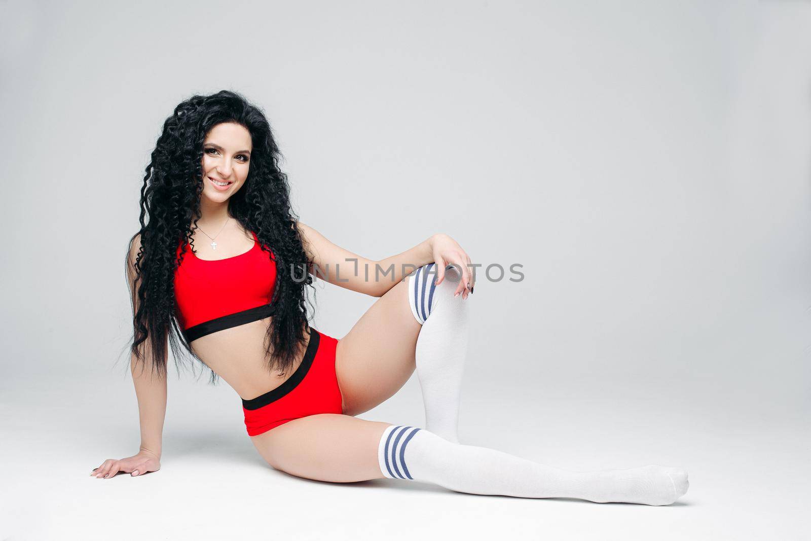 Full lenght portait of sexy dark haired girl wearing in red sports wear and white leggings lying on floor. Woman posing at white studio background, smiling at camera. Concept of sport, fitness, dance.