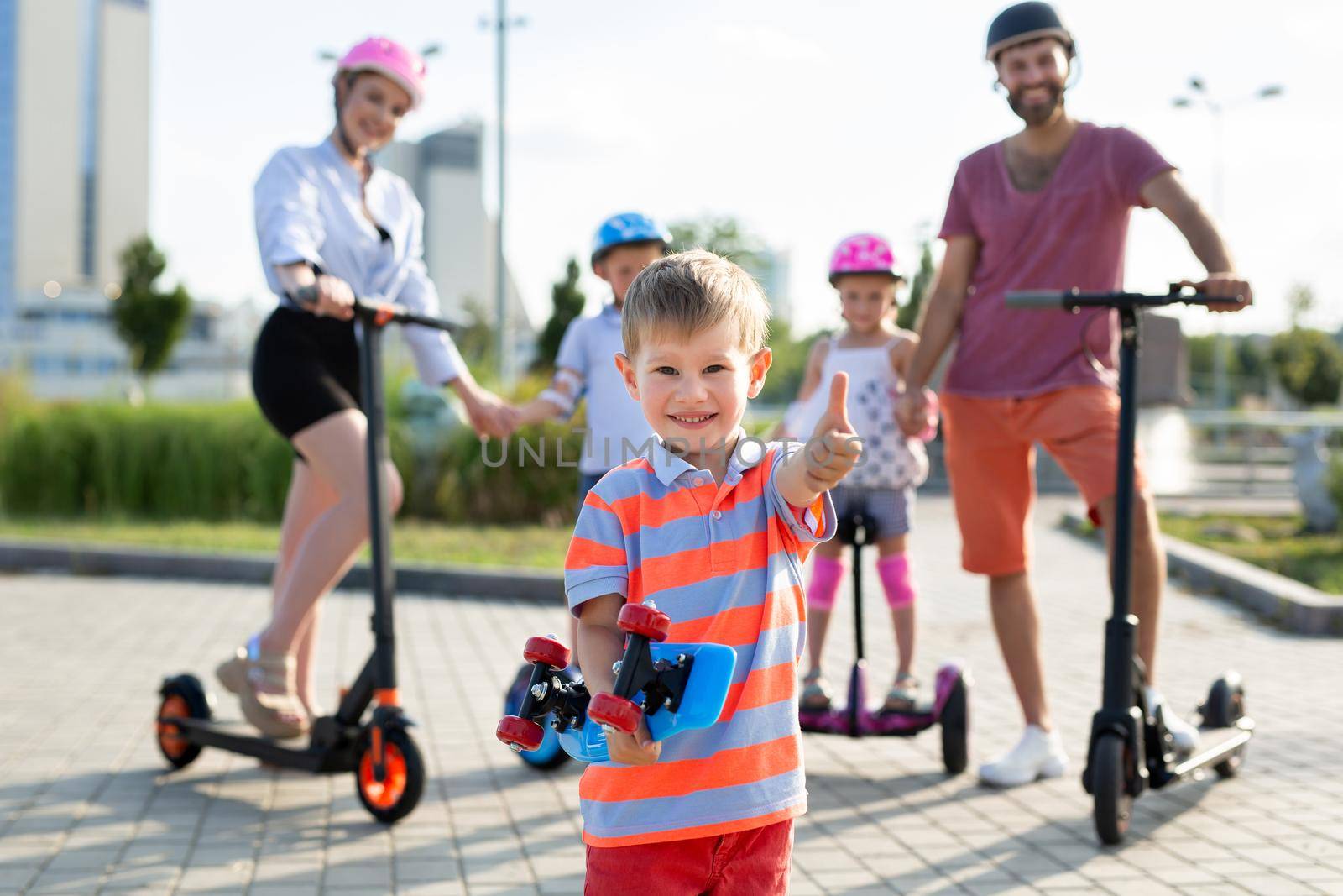 Happy family rides electric scooters and gyroscuters in the park, in the foreground a small boy holds a skate in his hands and gives a thumbs up