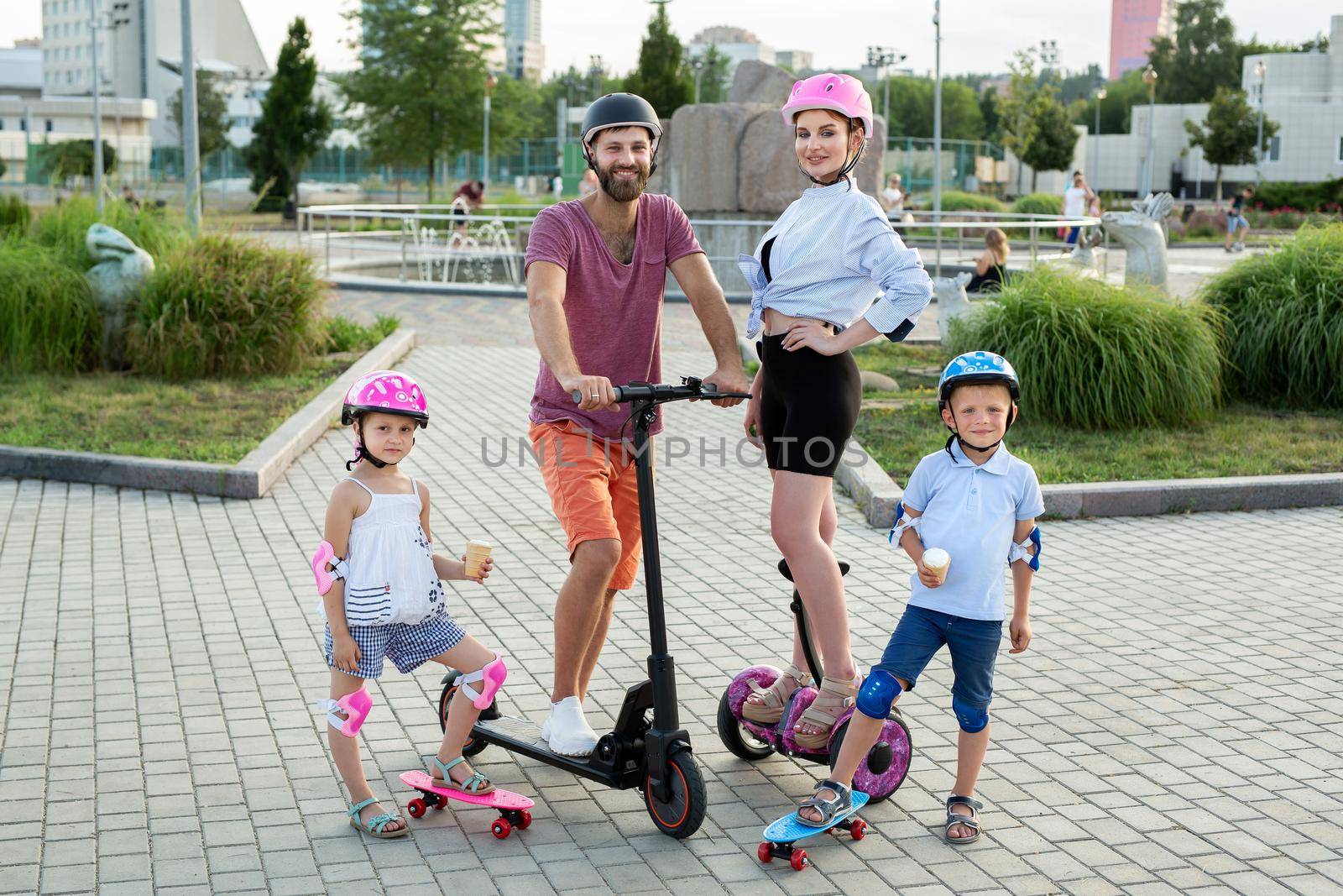 Happy family with kids riding on Segways, electric scooter and skateboards in the Park in the summer, children eating ice cream by StudioPeace