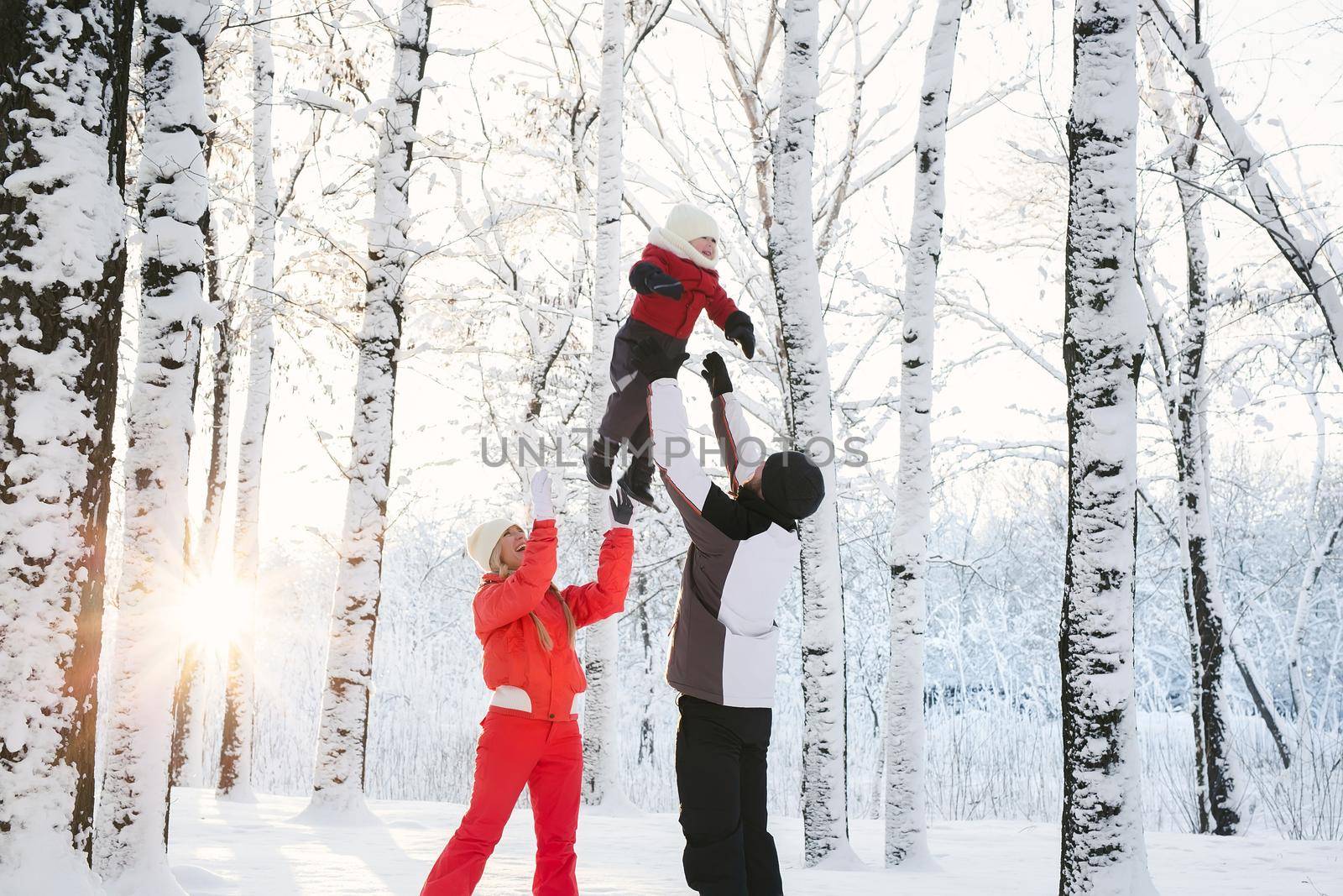 A young family walks in a snowy forest in winter, playing, laughing and throwing their son up.
