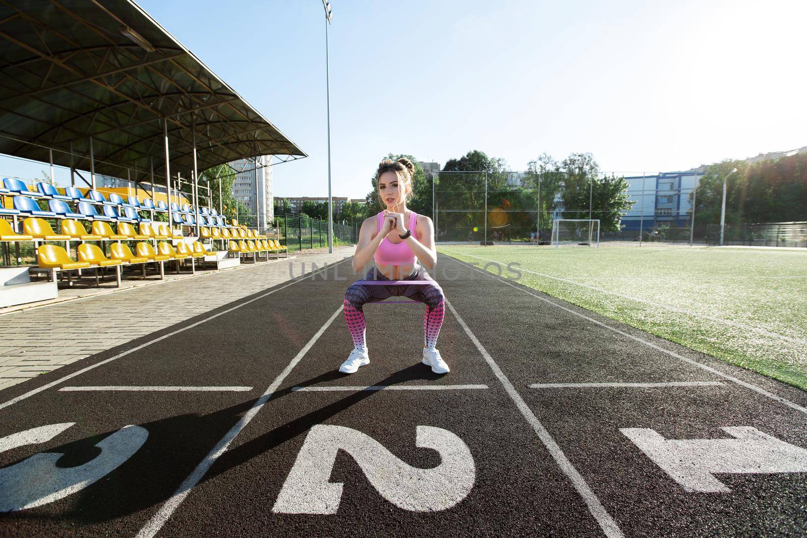 Woman squats with an elastic band and jumps up at the stadium in the summer. Sports lifestyle. by StudioPeace