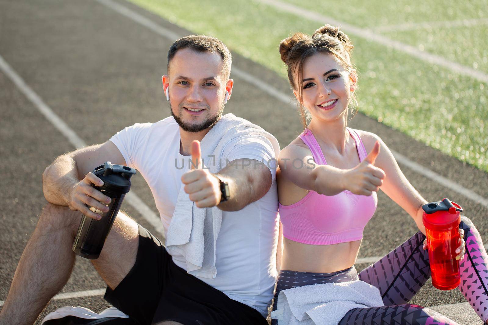 Young man and a girl sit on a treadmill in a stadium with water bottles and give a thumbs up. by StudioPeace