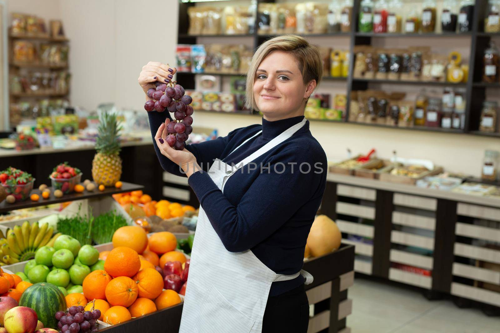 Female saleswoman helps a customer buy fruits and vegetables at a grocery store, holds them in her hands and shows them grapes.