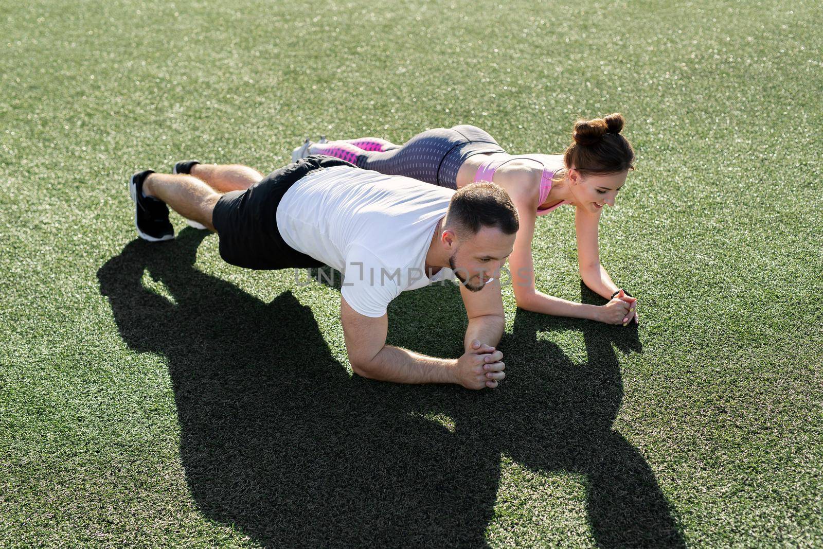 Close-up of a man and a woman doing a plank exercise on the grass at a sports stadium at sunset