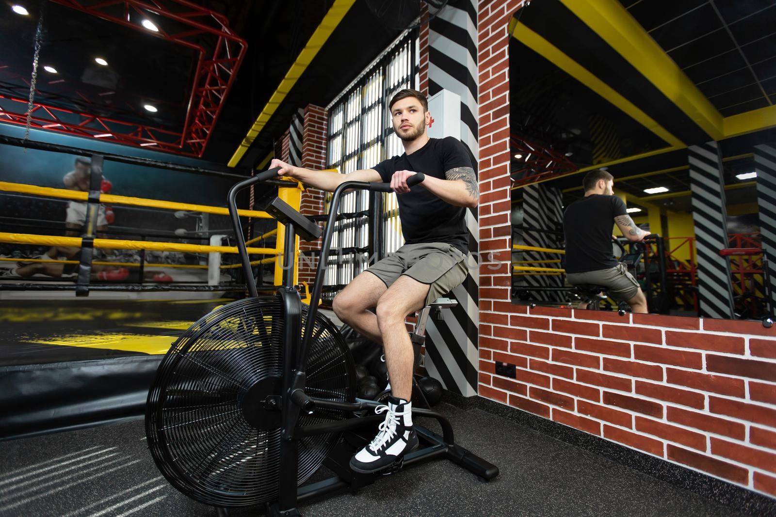 Young athletic male boxer trains on an exercise bike near the ring.