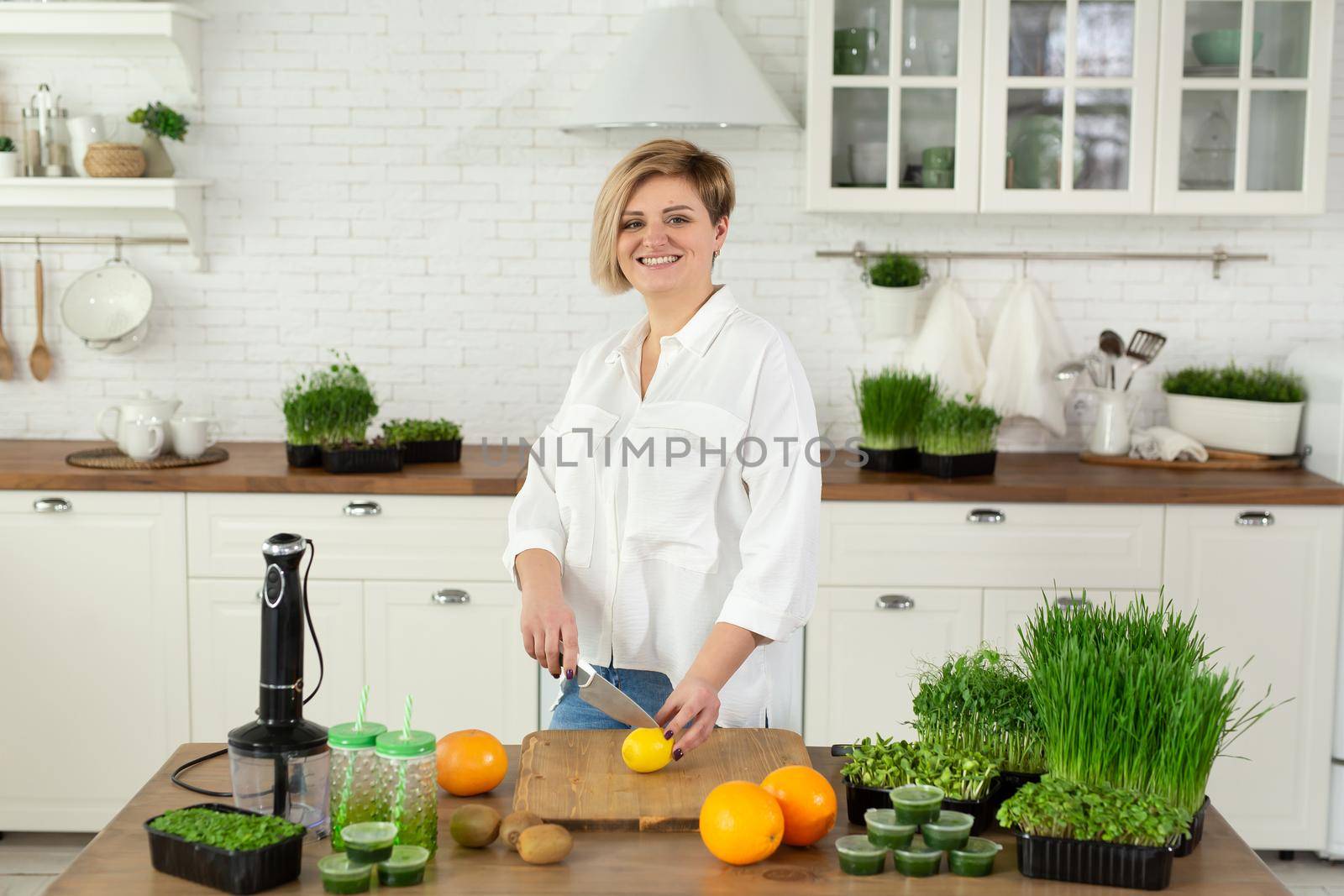 Young beautiful woman cuts a lemon in the kitchen on the table for a vitamin smoothie made of microgreen and fruit.
