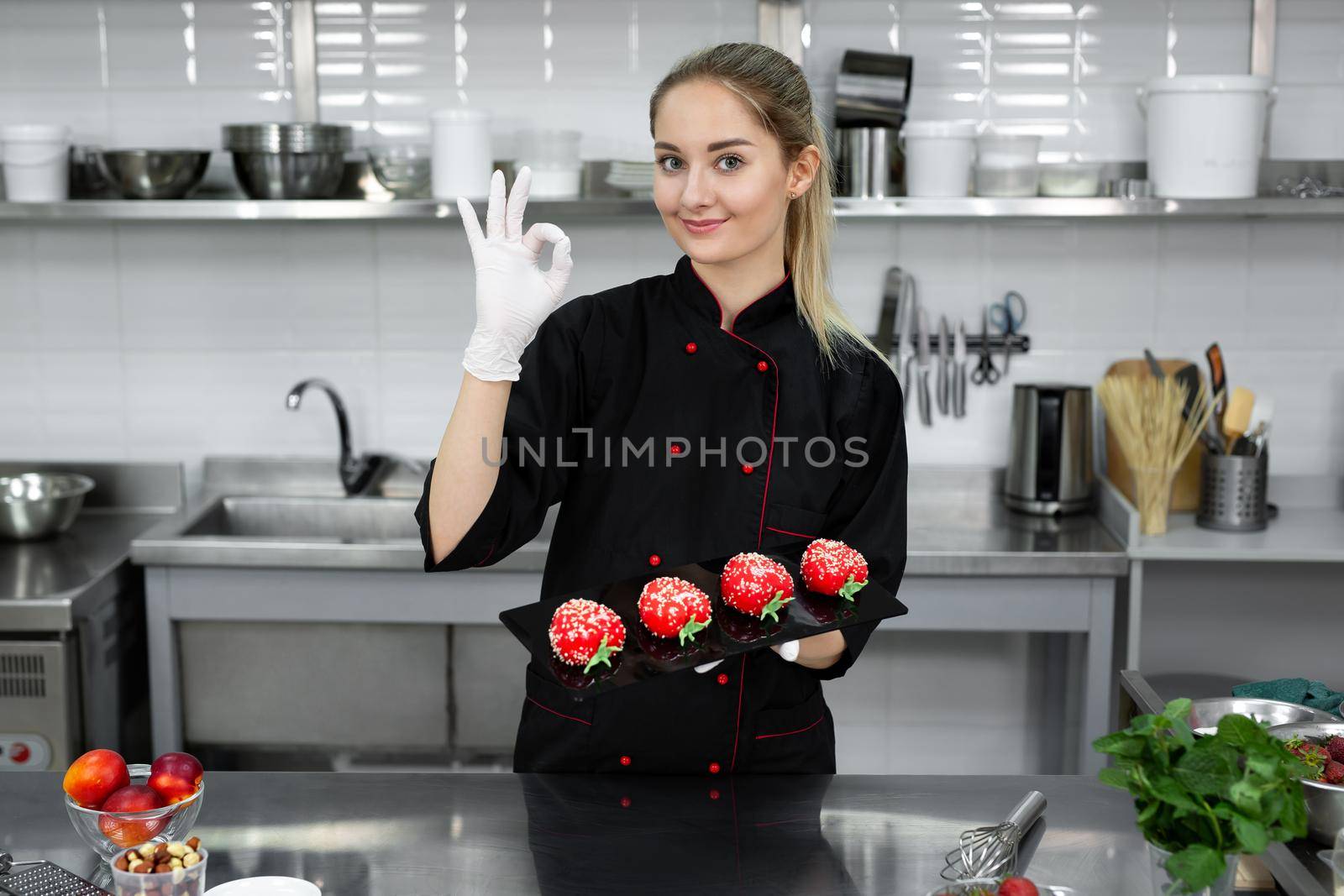 The pastry chef holds a tray with mass cakes, smiles and shows the sign ok