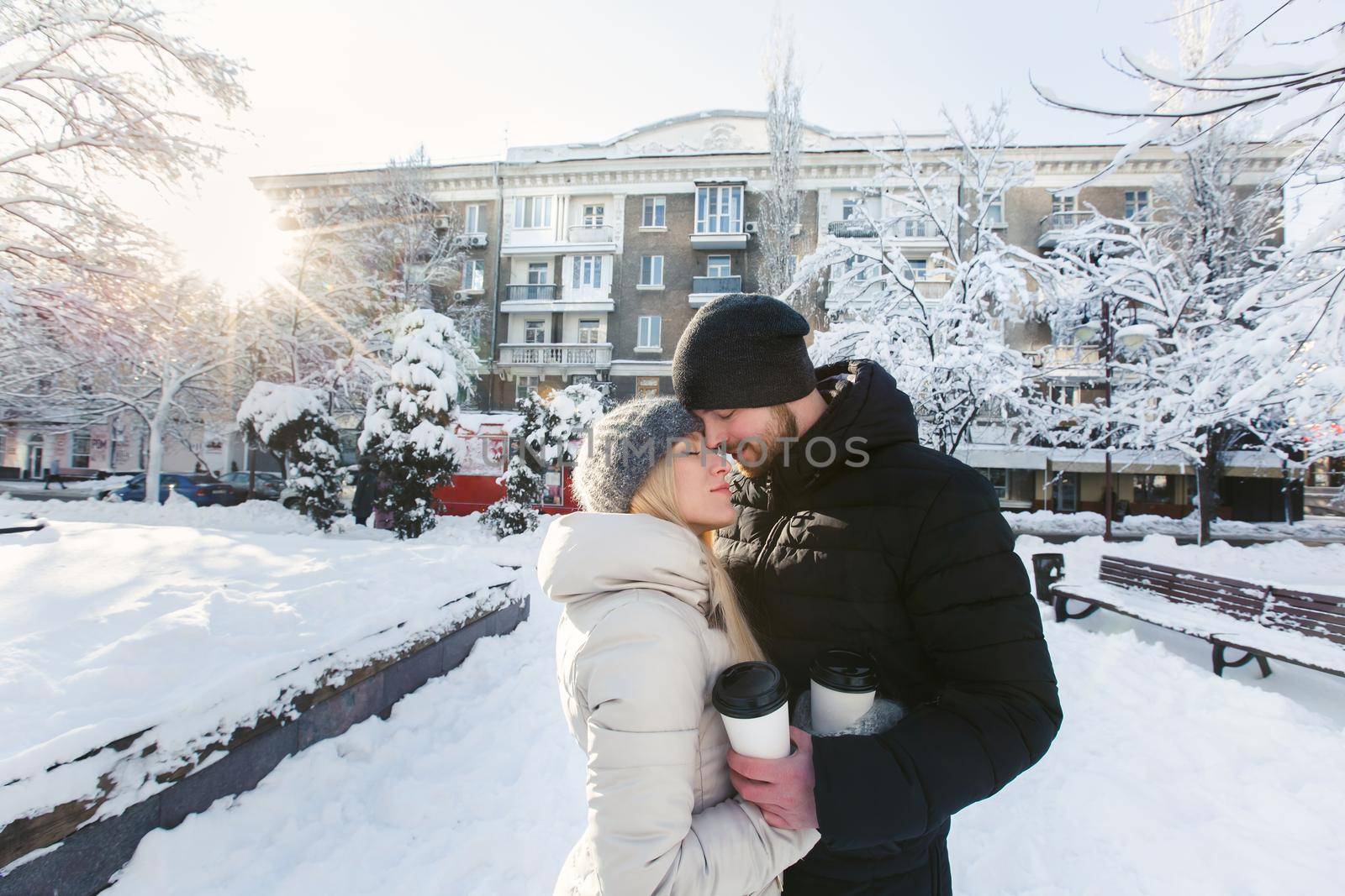 A loving beautiful couple in a winter snow park with coffee. A handsome man hugging his happy woman and laughing.