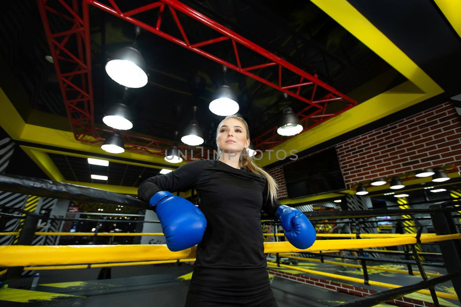 Portrait of a slender athlete in blue boxing gloves, leaning on a rope. A young female fighter poses near the boxing ring, looking away.