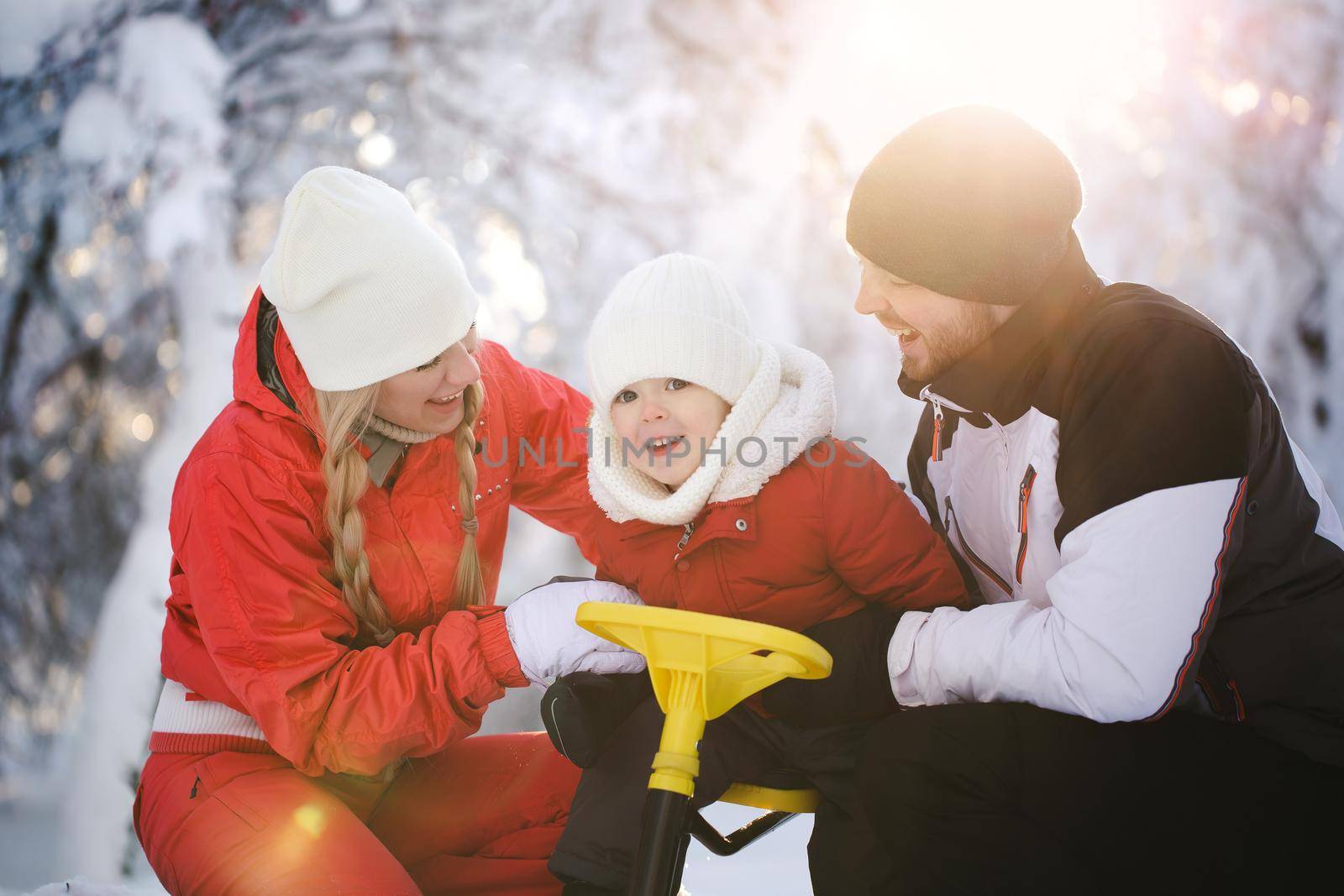 Portrait of a happy family in winter in the forest. The son is sitting on a sled, and dad and mom hug him and laugh.