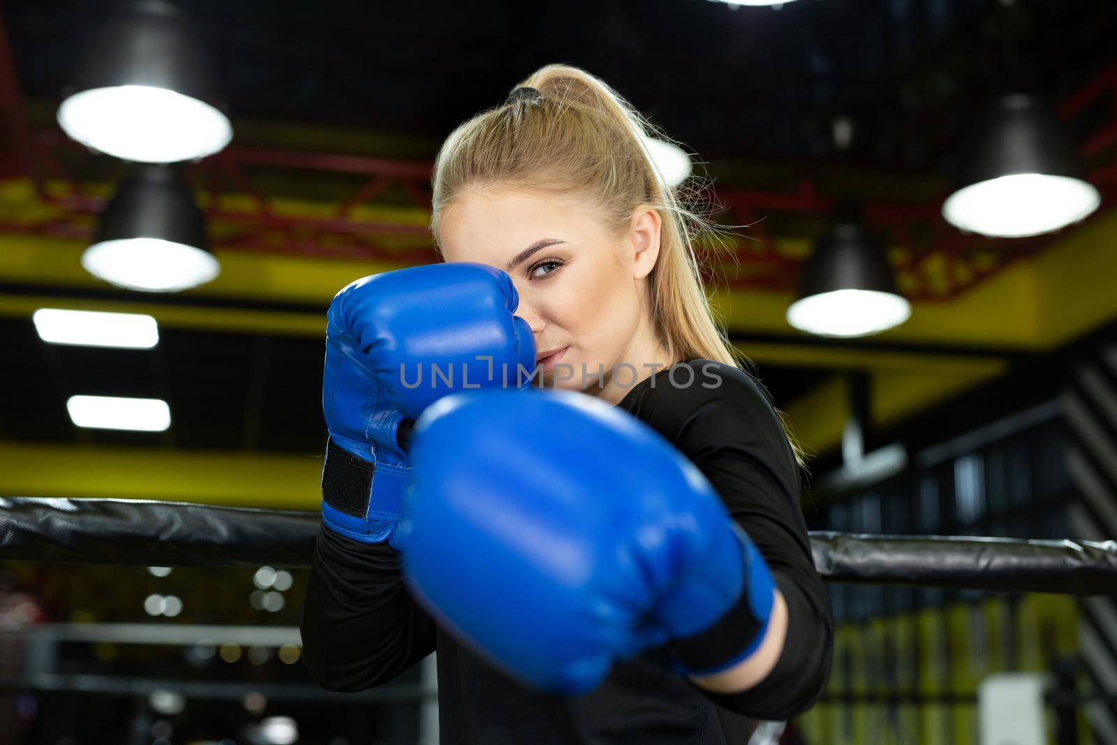 Young beautiful athlete in blue boxing gloves poses in the ring and looks at the camera by StudioPeace
