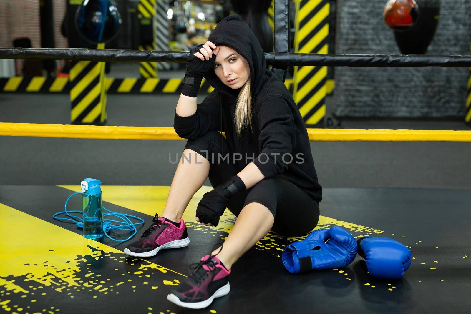 Young athletic girl in a black tracksuit sits in the ring and rests after a workout.