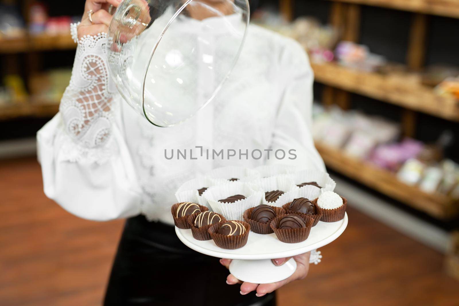 Handmade chocolates on a tray under a glass lid in the hands of a beautiful young woman in the store by StudioPeace