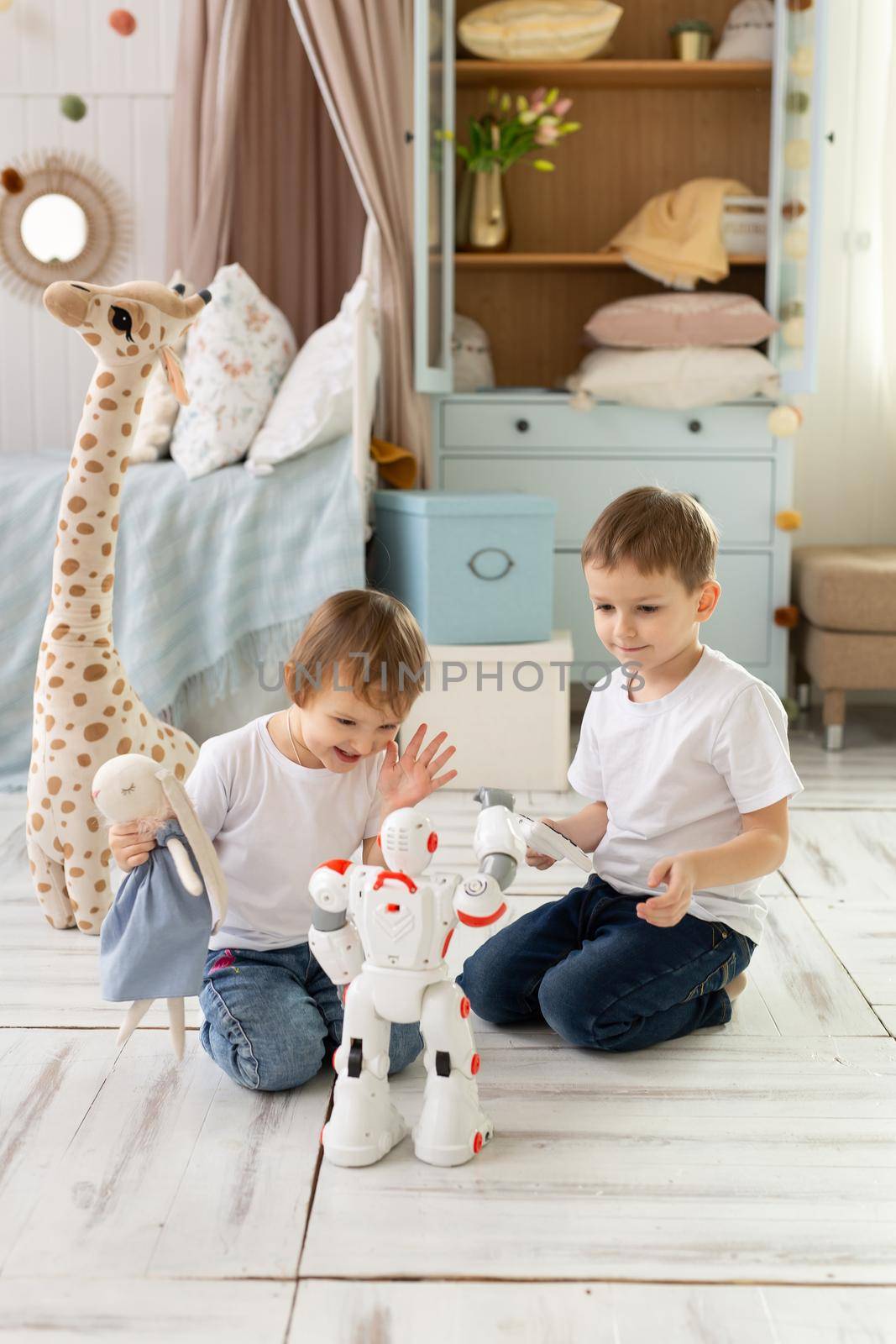 Small children brother and sister sit on the floor in the room, laughing and playing with the robot