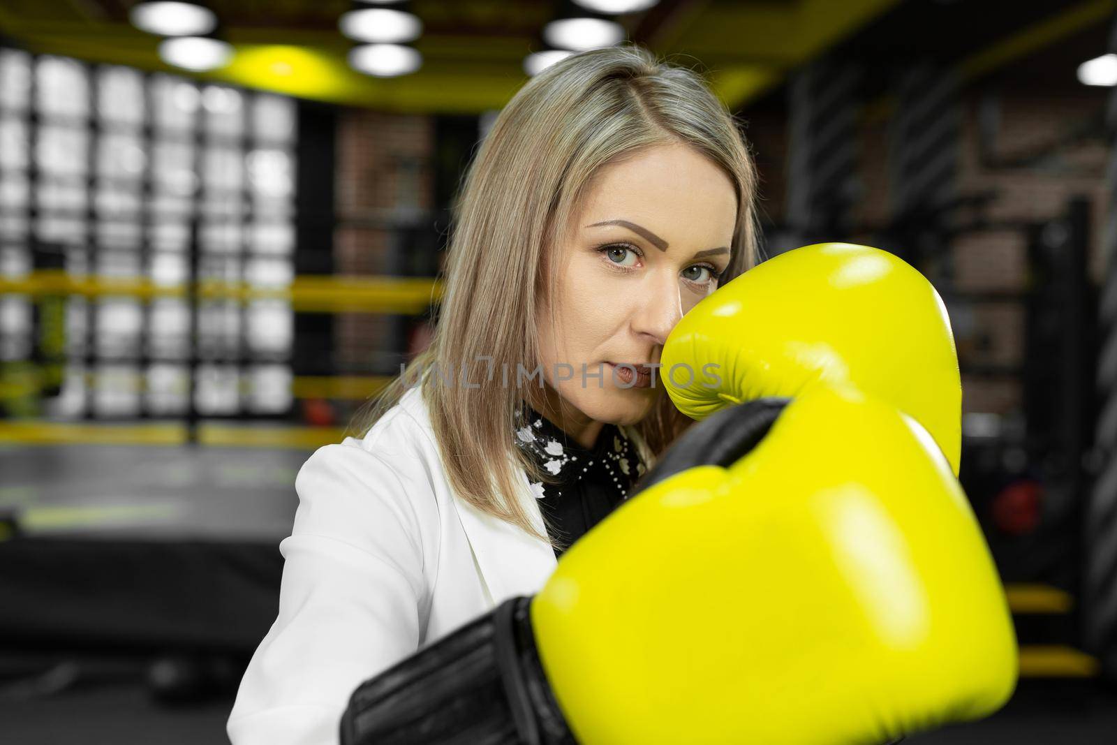 Determined, stylish businesswoman in yellow boxing gloves throws a punch at the camera against the backdrop of a boxing ring by StudioPeace