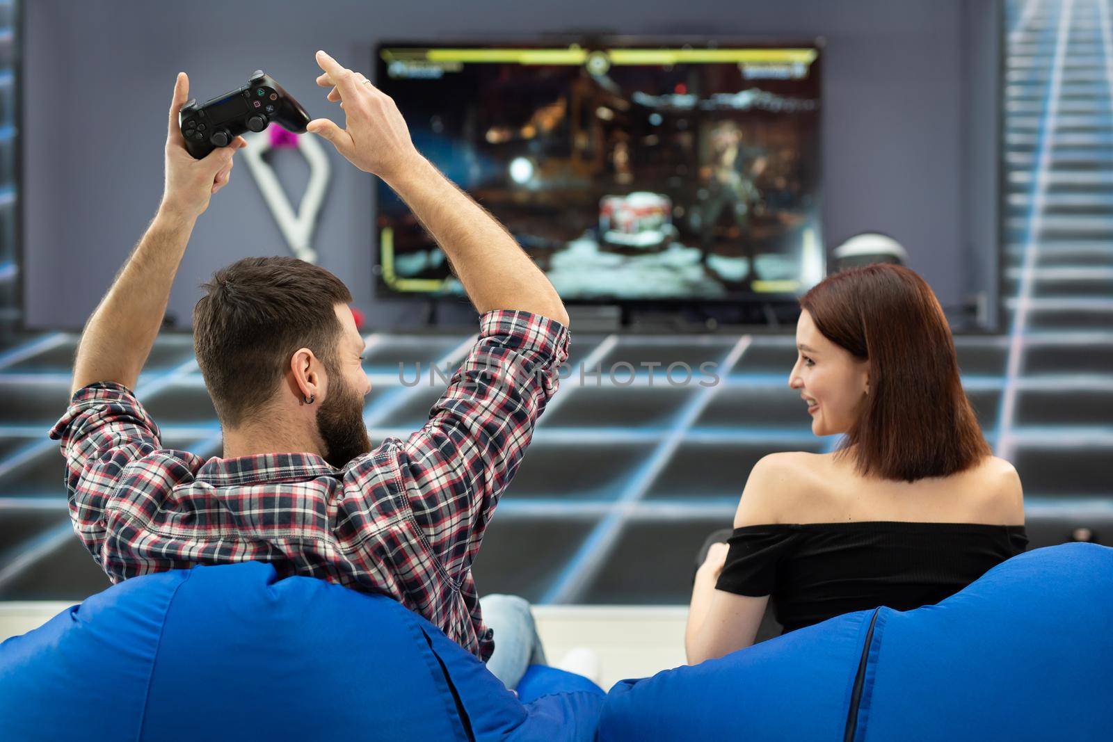 Young couple playing video games, sitting on chairs in a gaming club with controllers in their hands, a rear view from the TV screen.