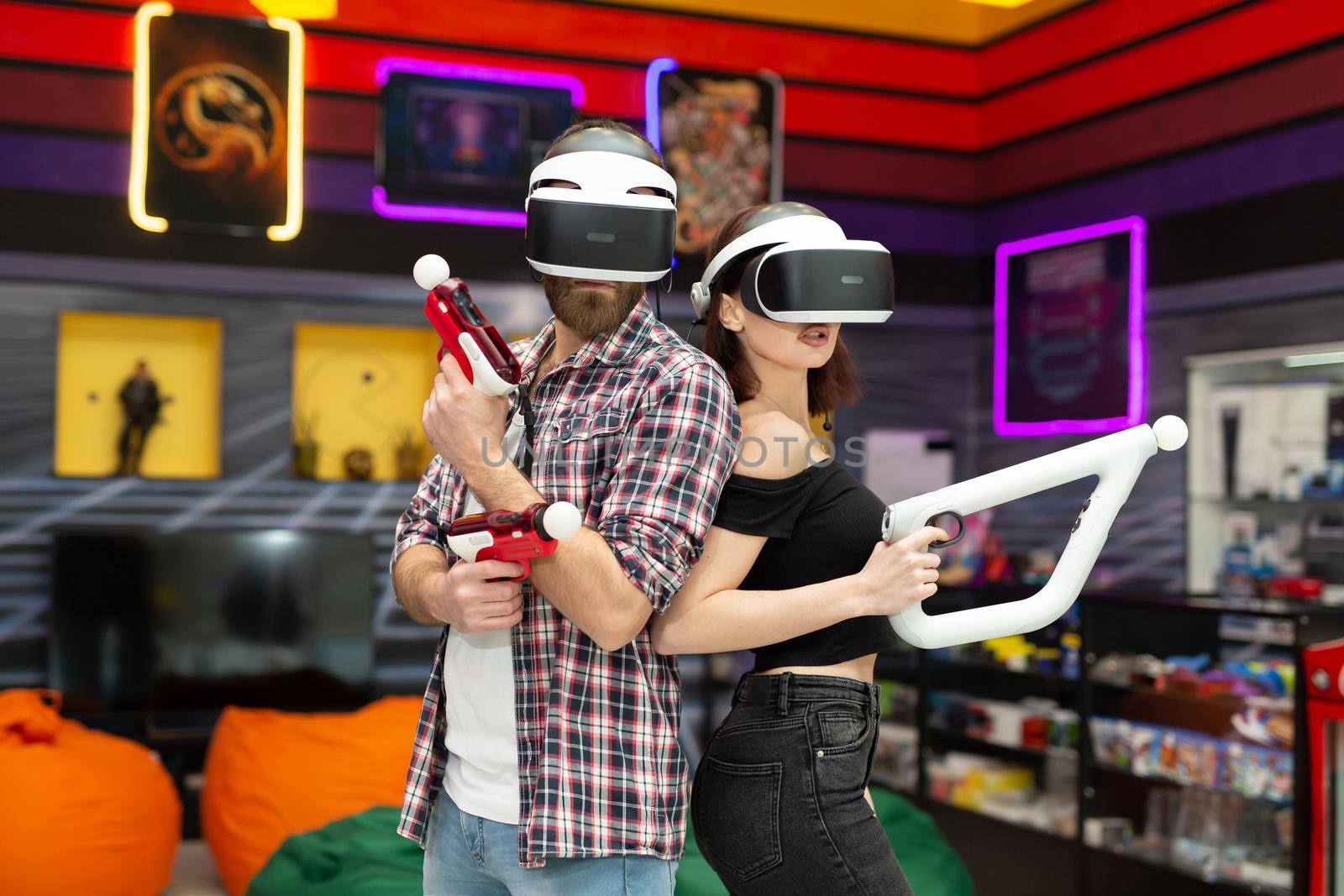 Friends, a man and a woman use a virtual reality headset with glasses and hand motion controllers and weapons in the play area.