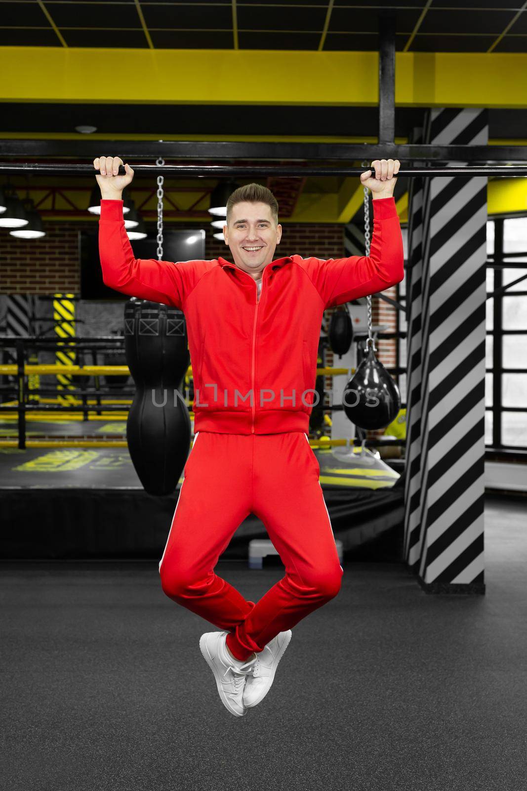 Thin man in a red tracksuit pulls himself up on a crossbar in the gym. by StudioPeace