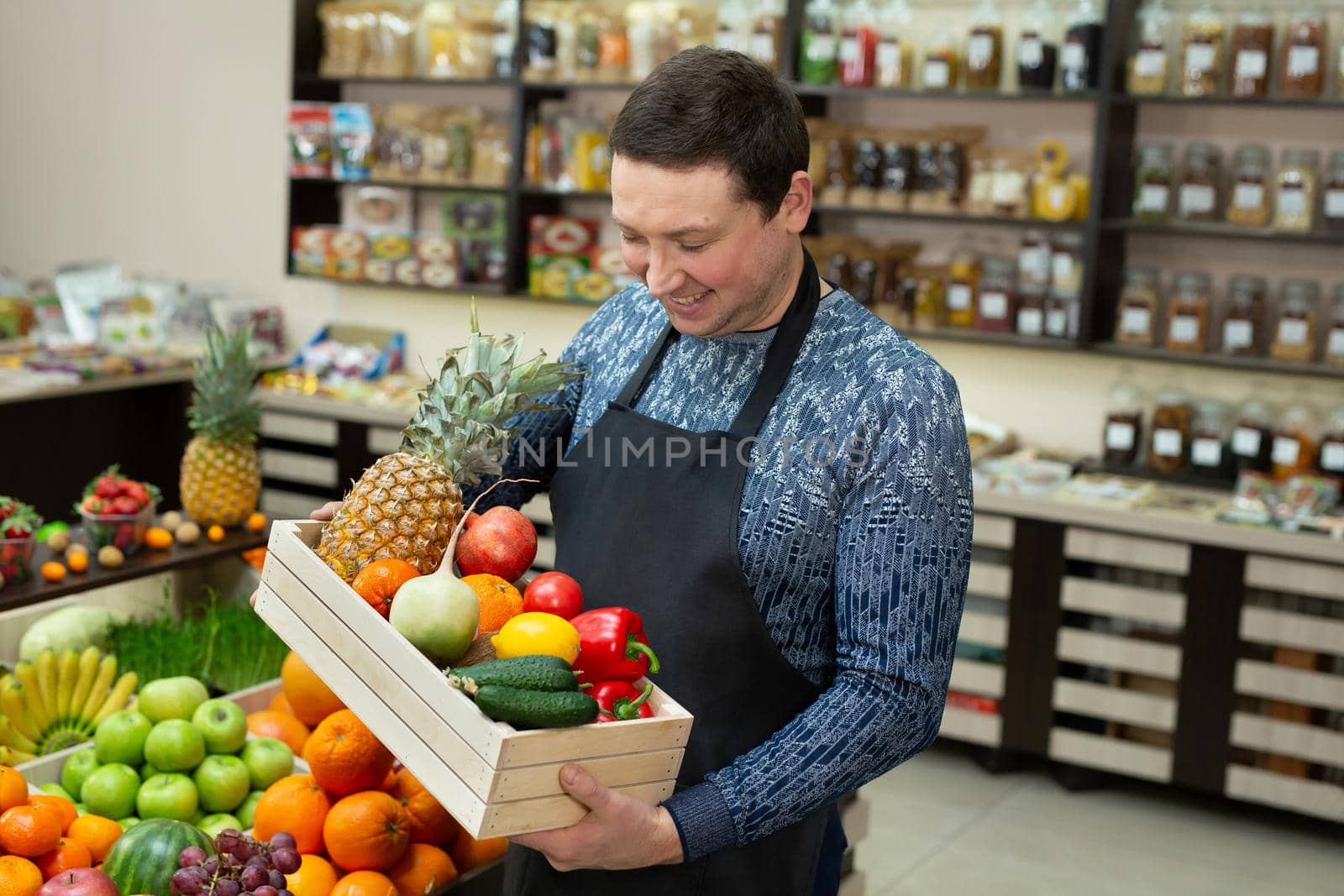 Smiling male salesman holds a wooden box with vegetables and fruits in the store by StudioPeace