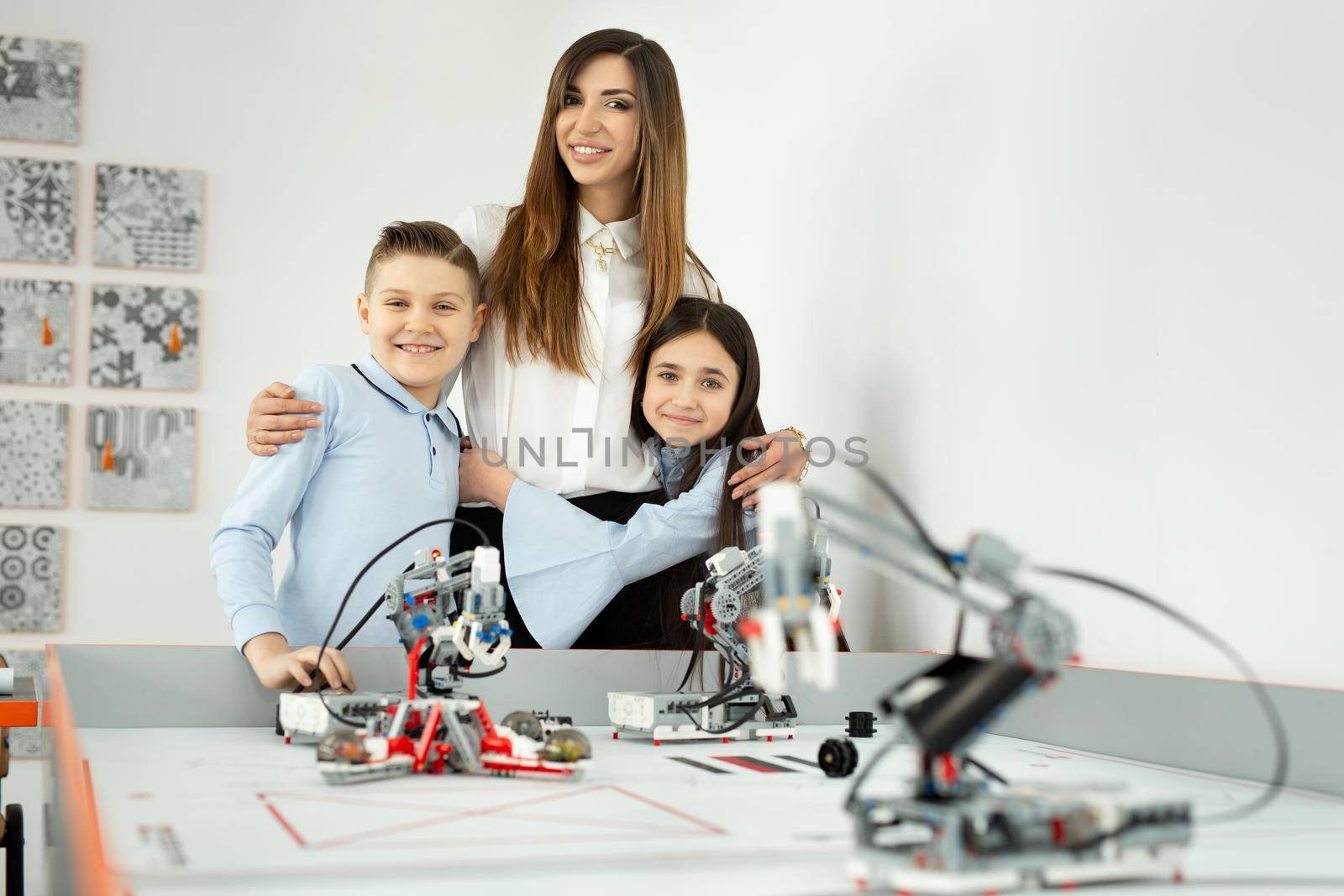 Young beautiful mother with her son and daughter pose at the school of robotics with robots from the constructor by StudioPeace