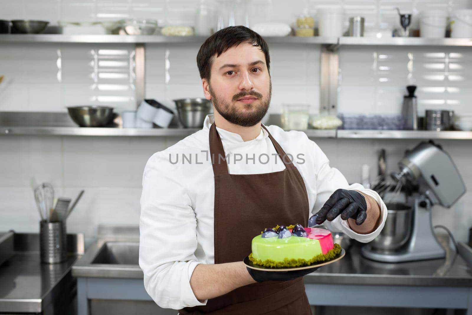 Young smiling chef holds a mousse cake decorated with handmade chocolates.