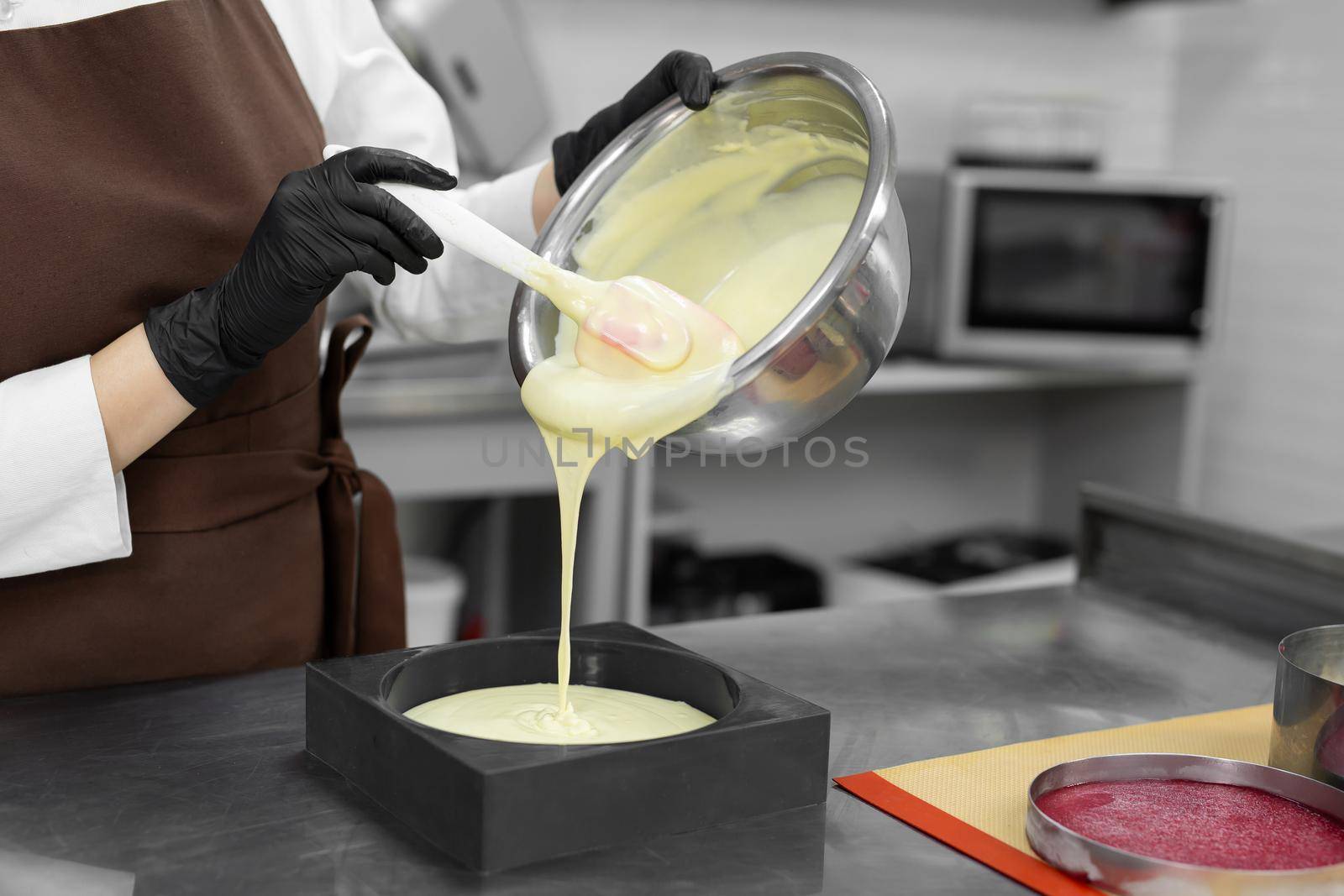 Young female pastry chef pours liquid mousse into a mold. Preparation of cake, dessert in a professional kitchen.