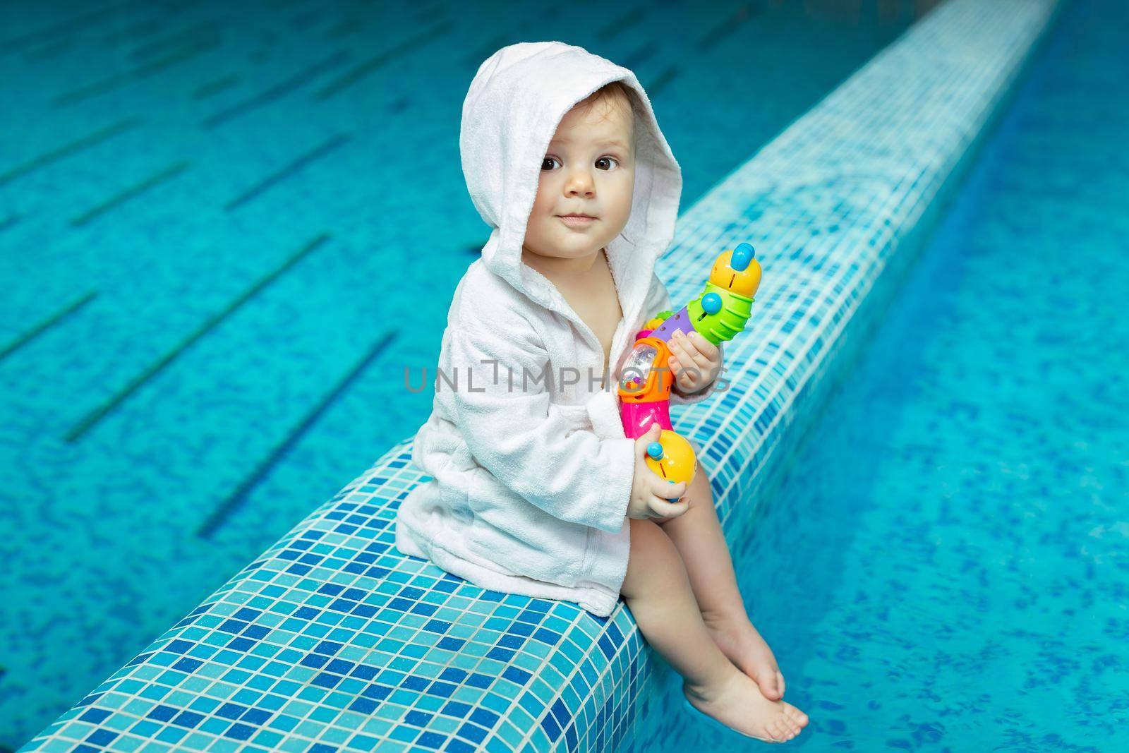 A toddler, a small child in a bath towel in the pool with a bright toy in his hands.