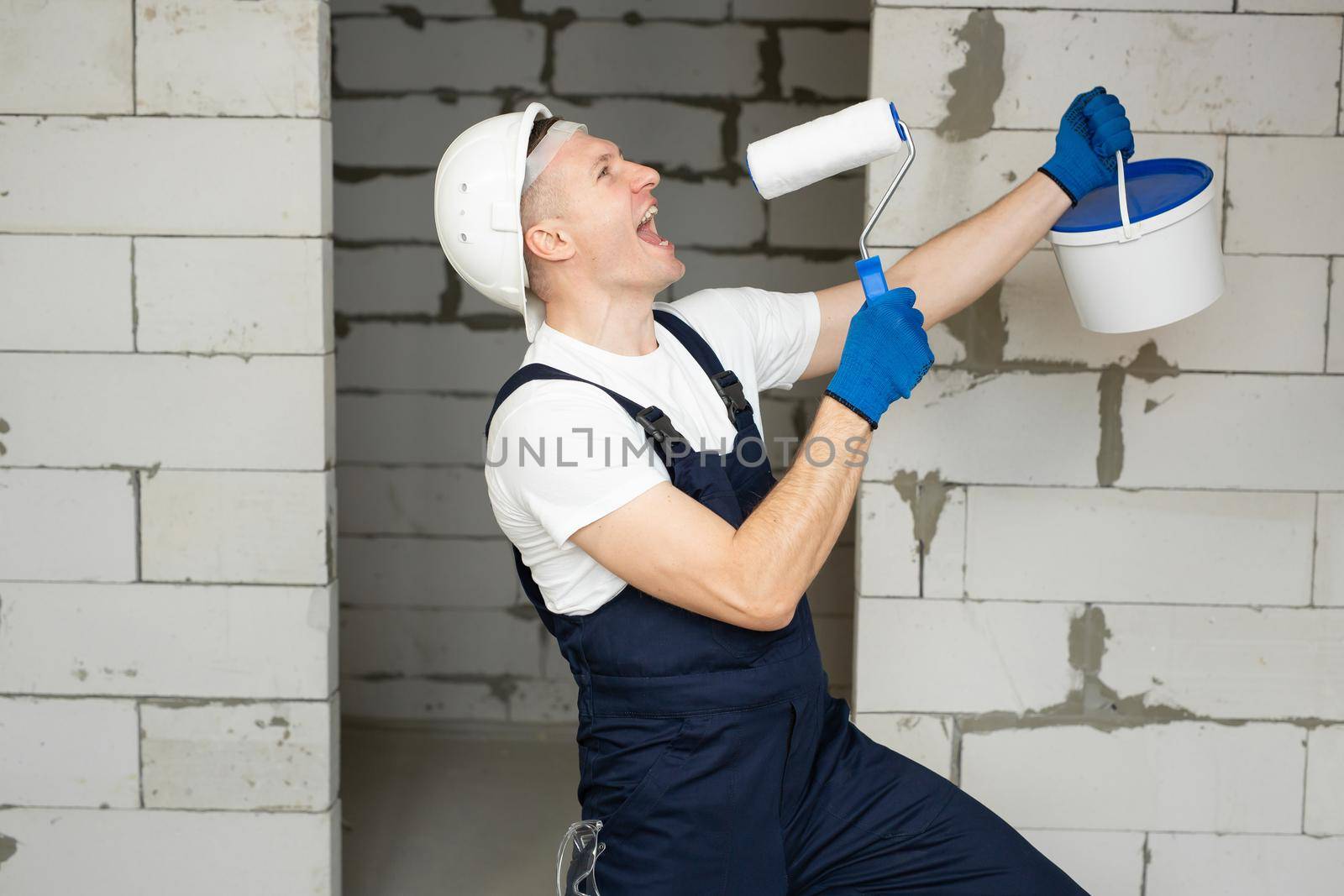 Male construction worker with paint and a roller sings and has fun on the construction site.