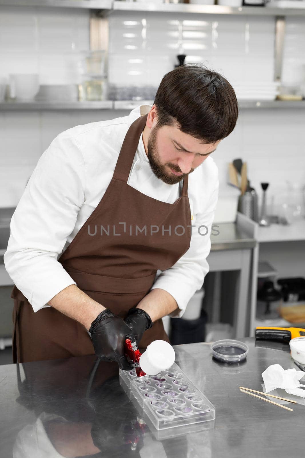 Male pastry chef holds chocolate candy molds and sprays food paint with a pastry airbrush in the kitchen by StudioPeace