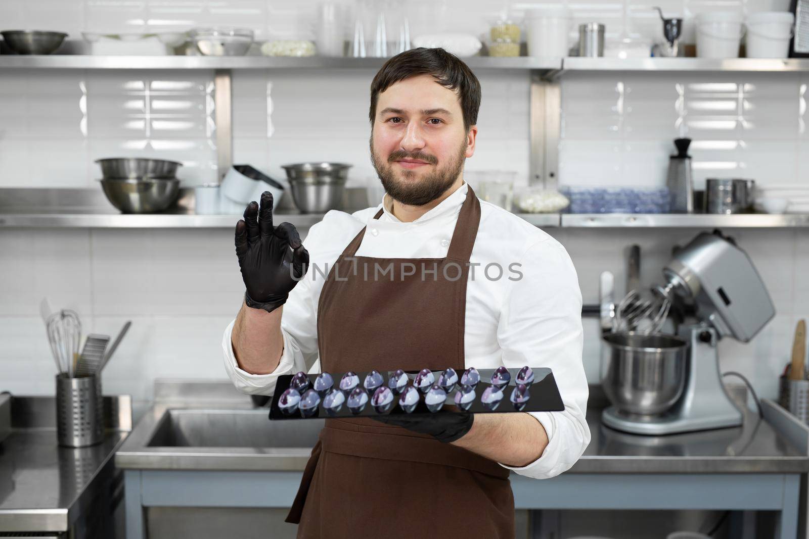 Male pastry chef in a professional kitchen holds handmade chocolates in his hands and shows the ok sign.