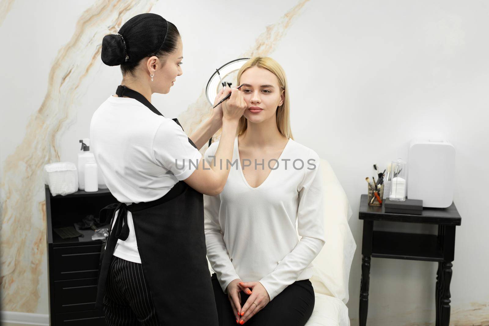 Make-up artist cosmetologist paints the eyes of a young beautiful woman in the salon with a pencil.