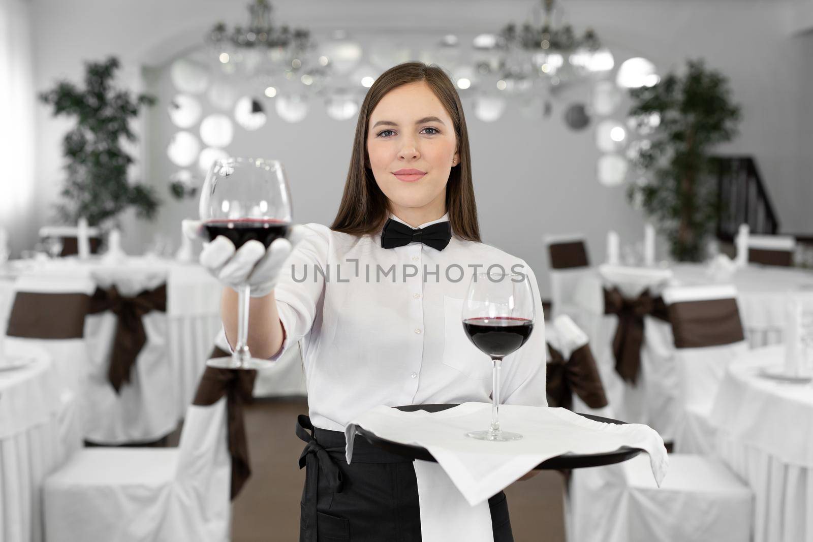 Female waiter in white gloves holds a glass and a bottle of red wine by StudioPeace