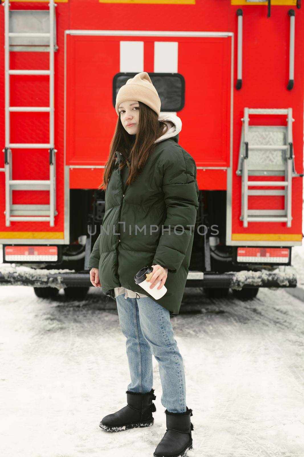 Young beautiful woman drinks coffee in winter on a city street with a red fire truck on the background.