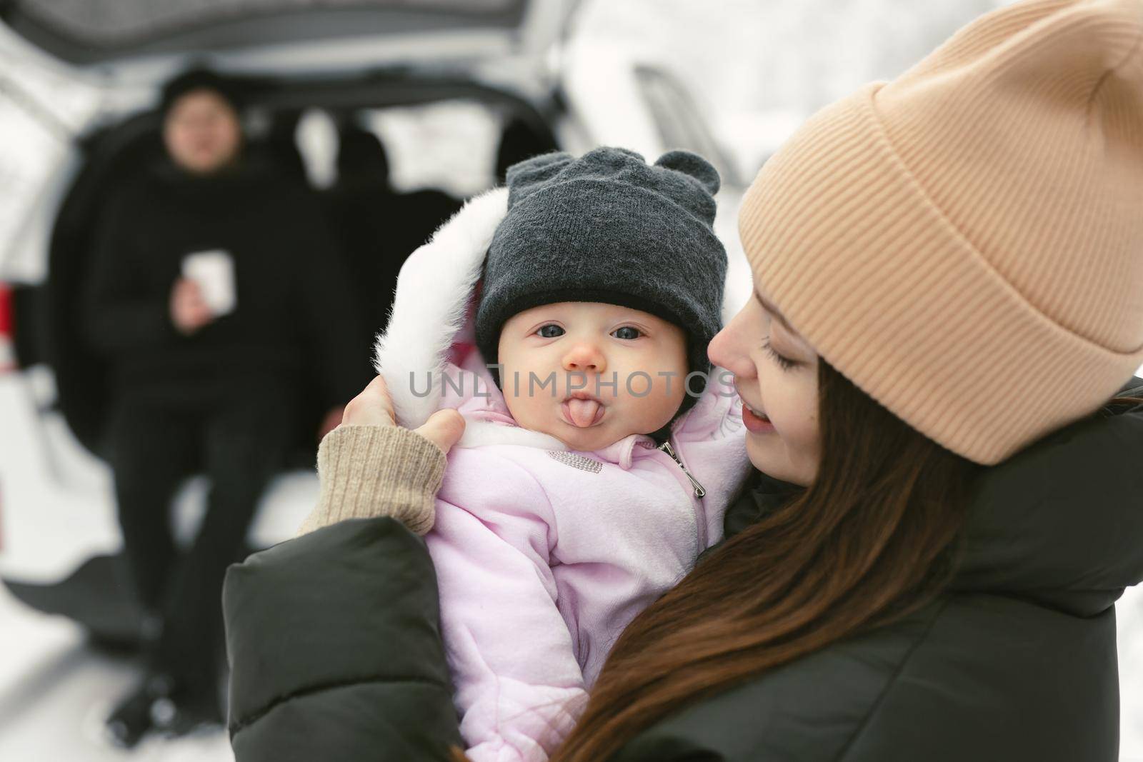 Baby girl looks into the camera and shows her tongue in her mother's arms in a winter snow-covered forest by StudioPeace