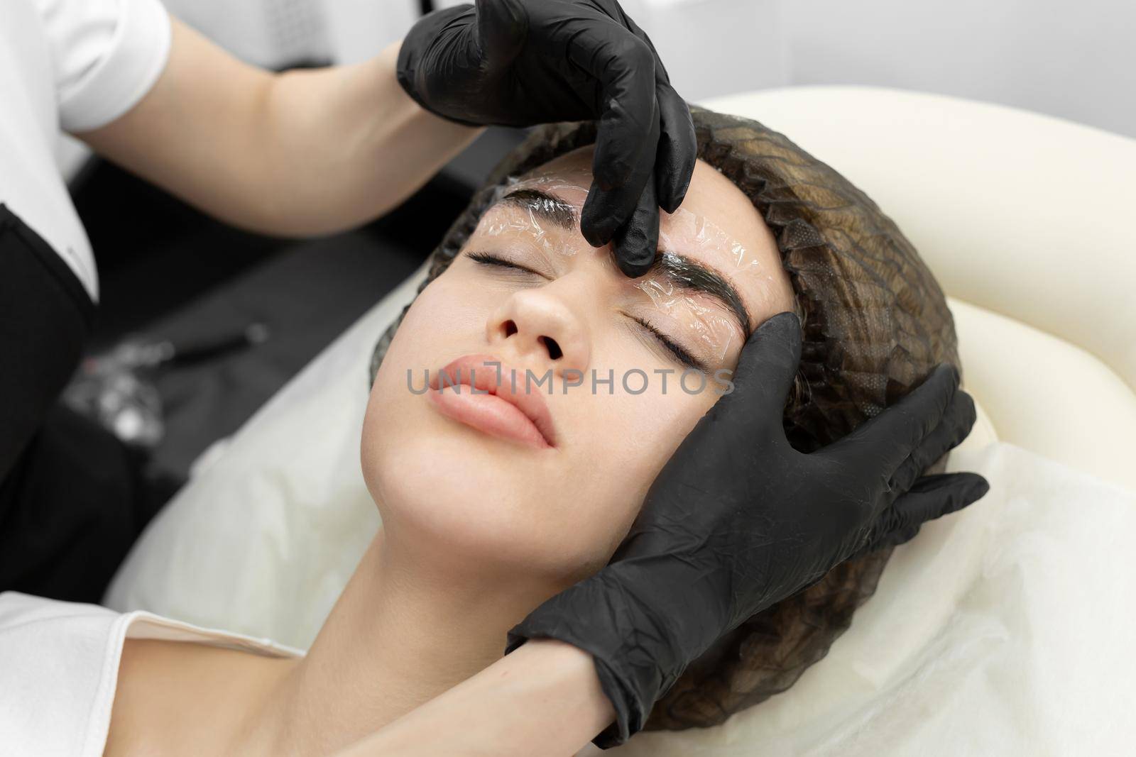 Girl's face with closed eyes close-up. A white cream is applied on the eyebrows - an analgesic and the cosmetologist's hands cover the eyebrows with a transparent film by StudioPeace