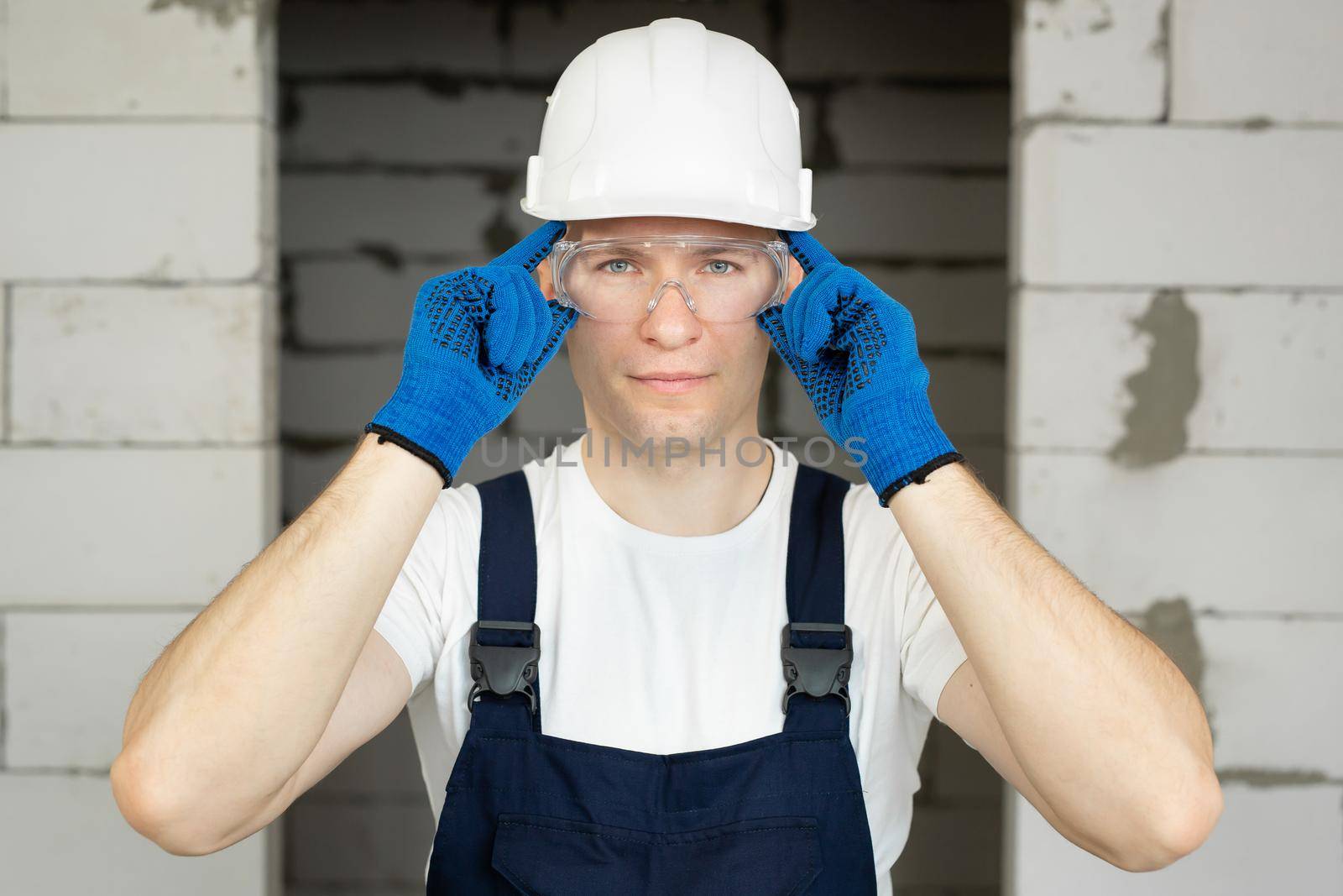 Handyman in a white T-shirt, a combination jacket and a hard hat puts on safety glasses.